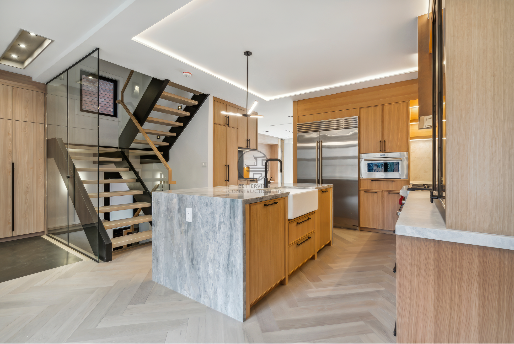 Modern open-concept kitchen with marble island, wood cabinetry, LED ceiling lighting, and adjacent floating staircase with glass railing.