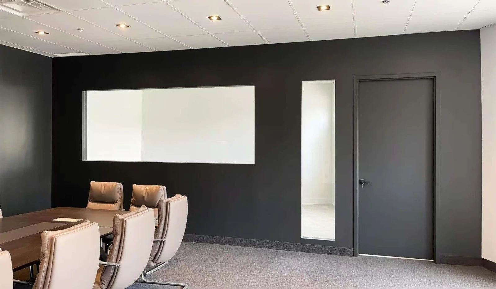 Modern conference room with black accent wall, beige chairs around a large wooden table, a long rectangular window on the wall, and a closed door to the right.