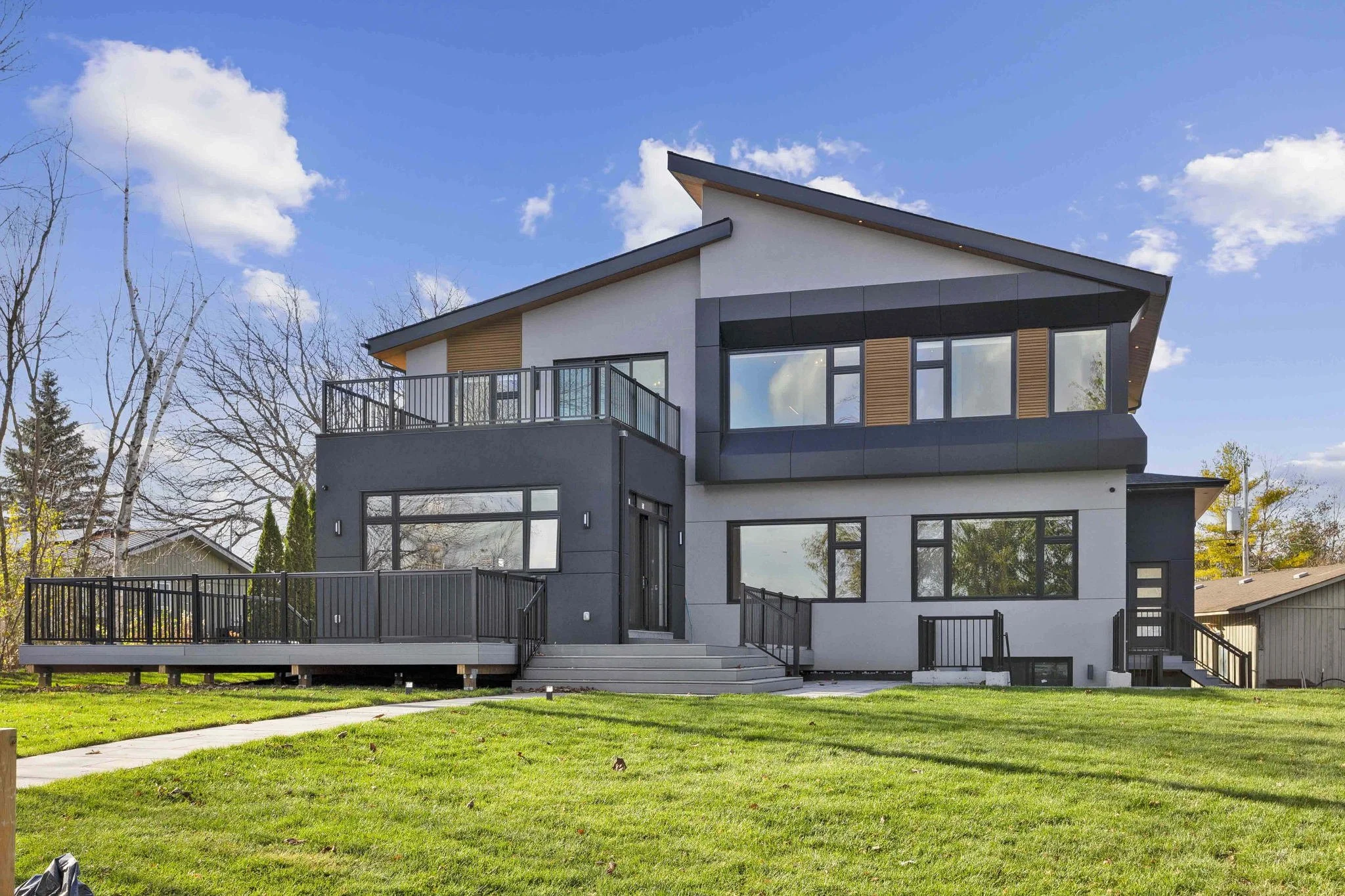 Modern multi-level house with large windows, black and white exterior, and a balcony, surrounded by a green lawn and trees with a blue sky and clouds above.