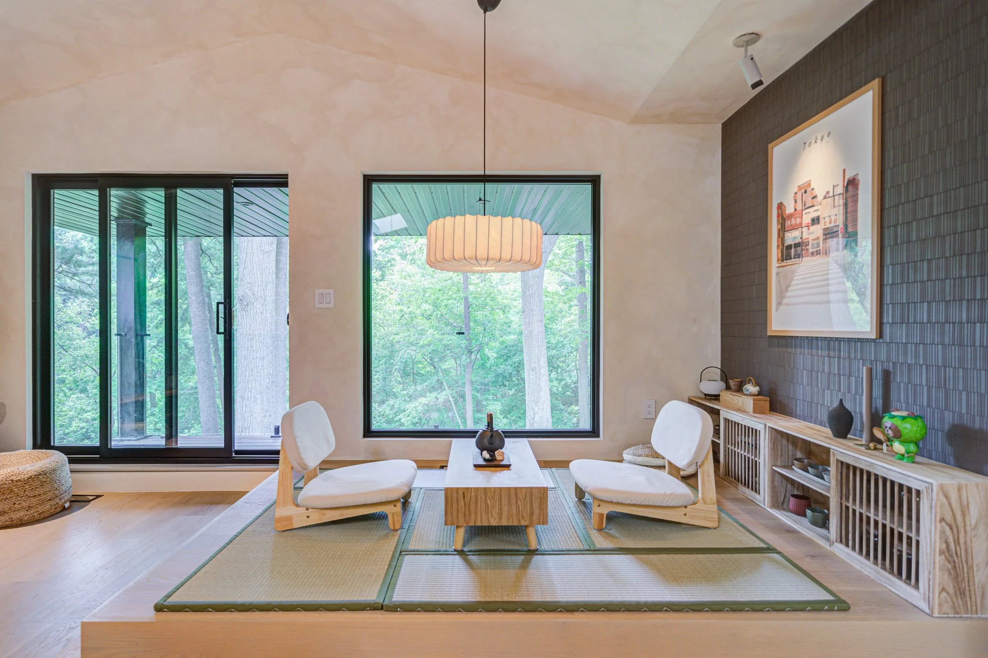 A dining area with a wooden table and four white cushioned chairs on a green tatami mat, large windows showing a green outdoor view, and modern decor including a hanging paper lantern, framed artwork, and a sideboard with decorative items.