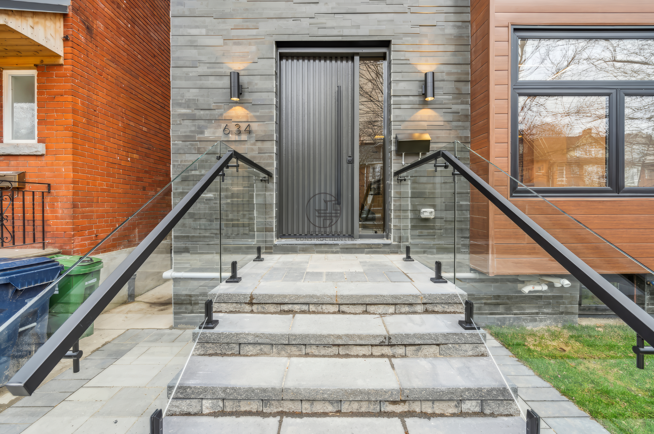 Modern home entrance with stone steps, glass railing, contemporary front door, and exterior wall lighting with mixed brick and stone facade.
