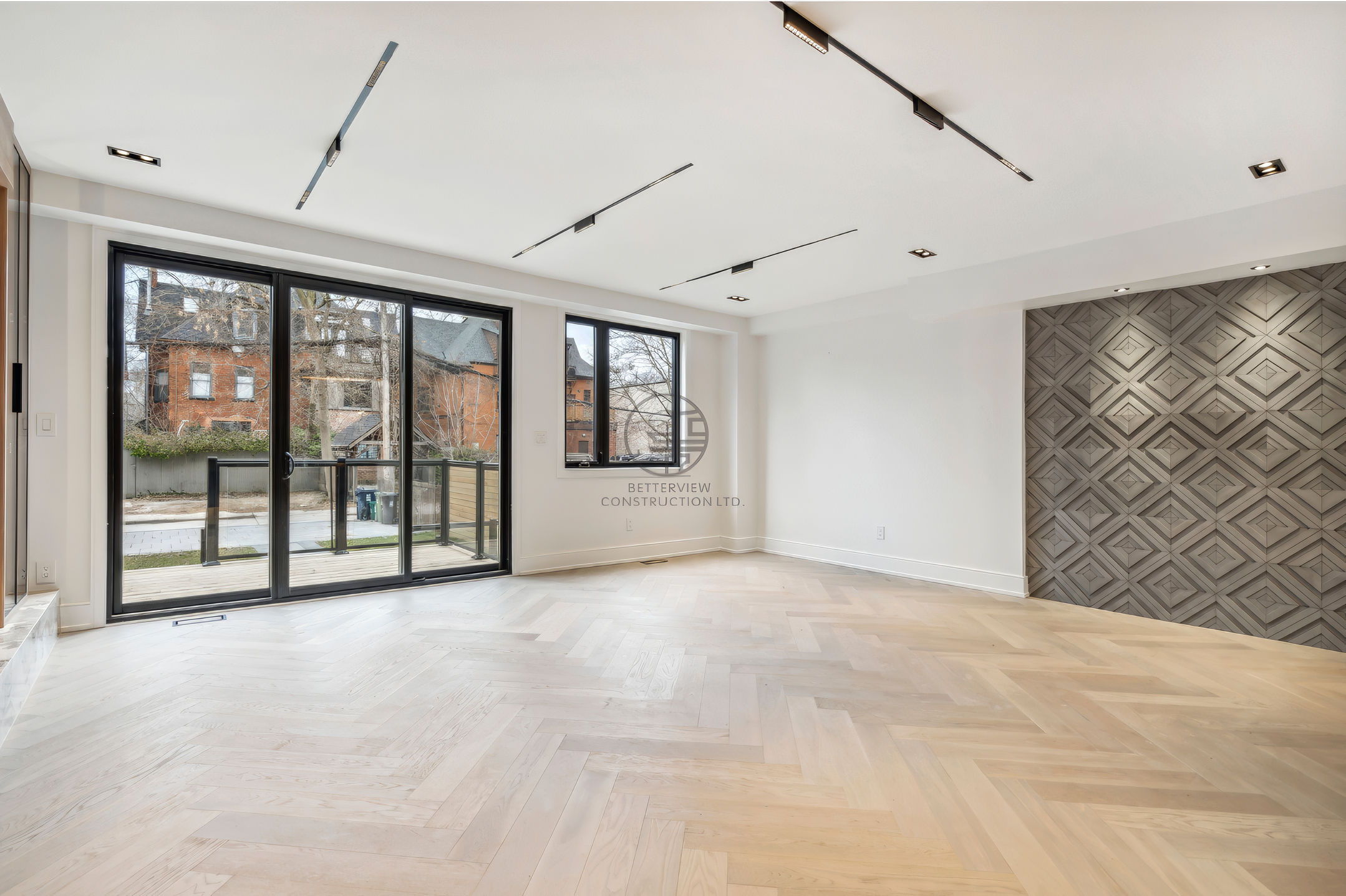 Spacious open living room with herringbone wood flooring, large sliding glass doors, geometric feature wall, and modern recessed lighting.