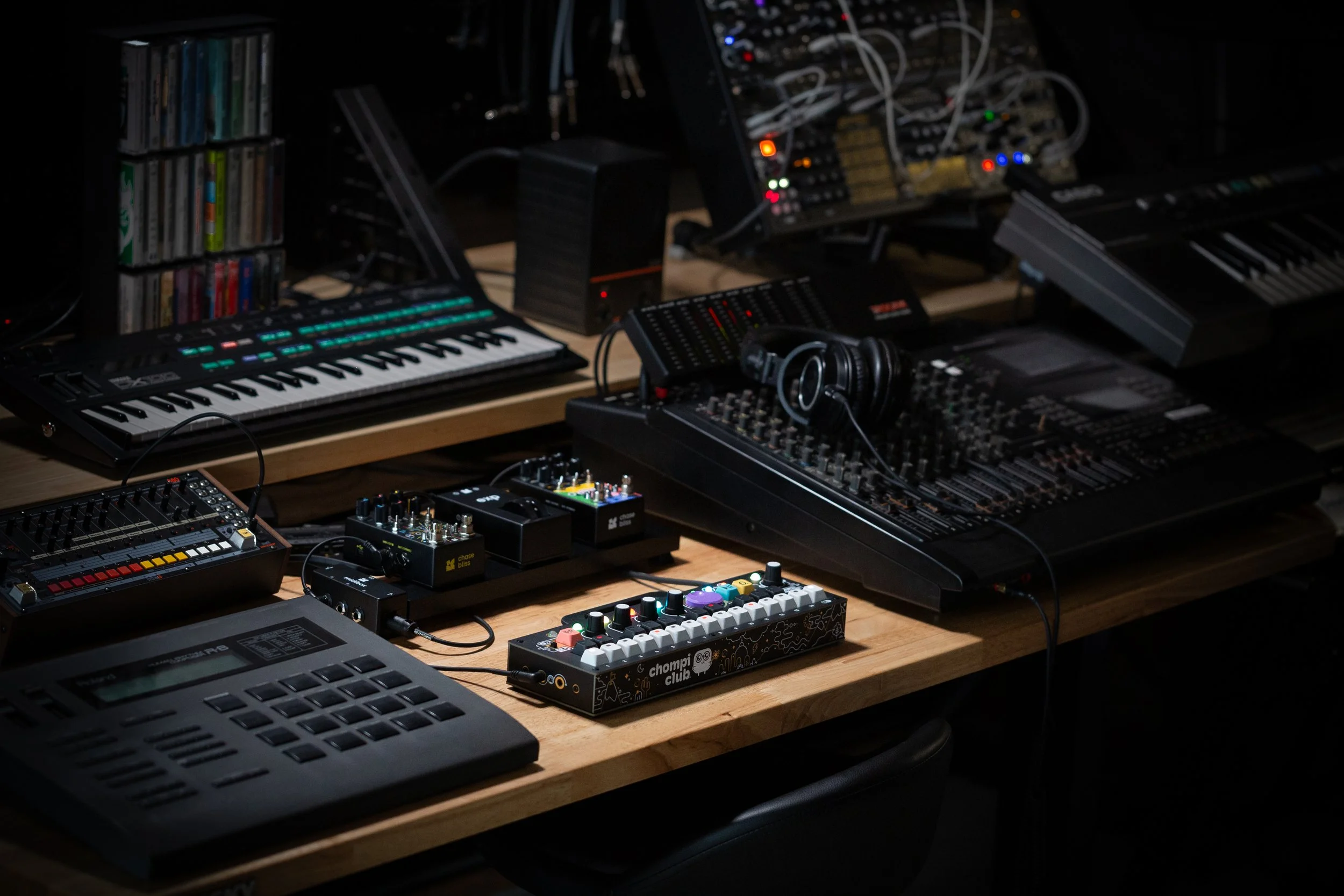 CHOMPI on a  table surrounded by a variety of synths and musical equipment.
