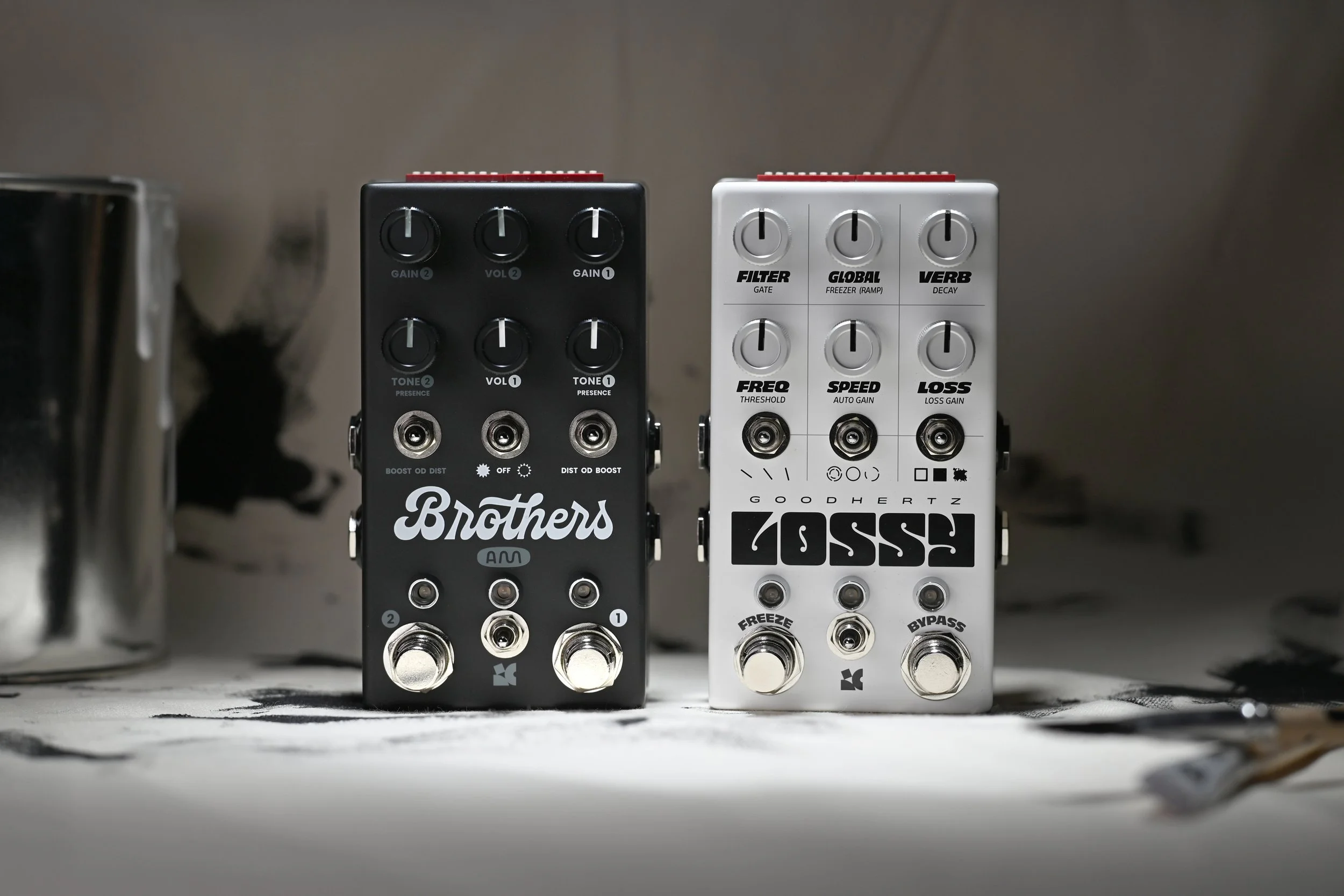 Brothers AM and Lossy Monochrome limited edition colorway pedals sitting on a table covered with paint and paintbrushes.