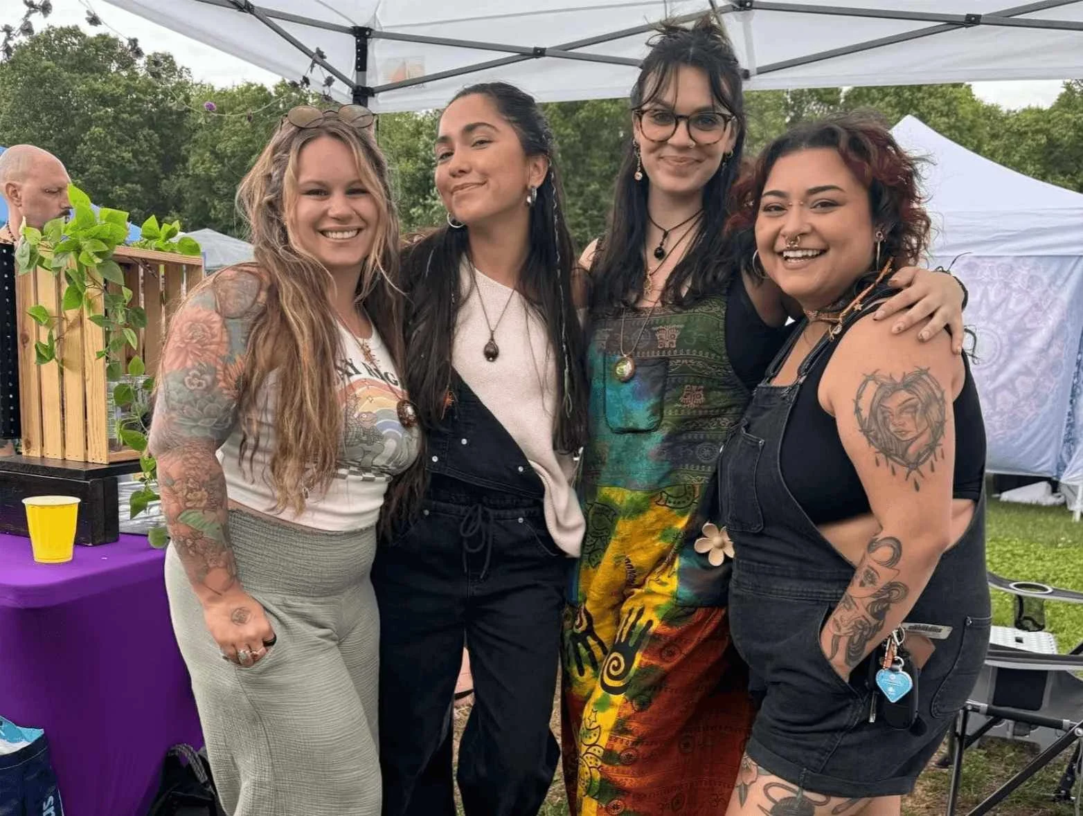 Four women standing together at an outdoor event, smiling, with tents in the background. They are dressed casually with colorful tattoos visible on their arms and shoulders.