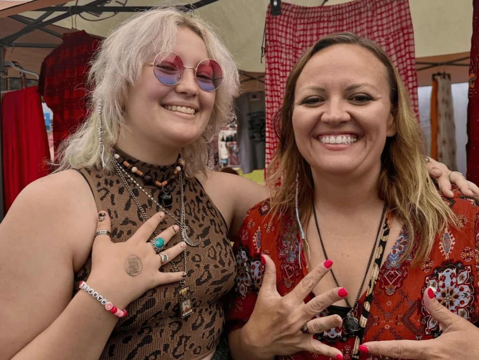 Two women smiling at a festival or outdoor market, making hand gestures. The woman on the left has curly blonde hair, wearing pink sunglasses, a sleeveless animal print top, and multiple necklaces and rings. The woman on the right has wavy blonde hai