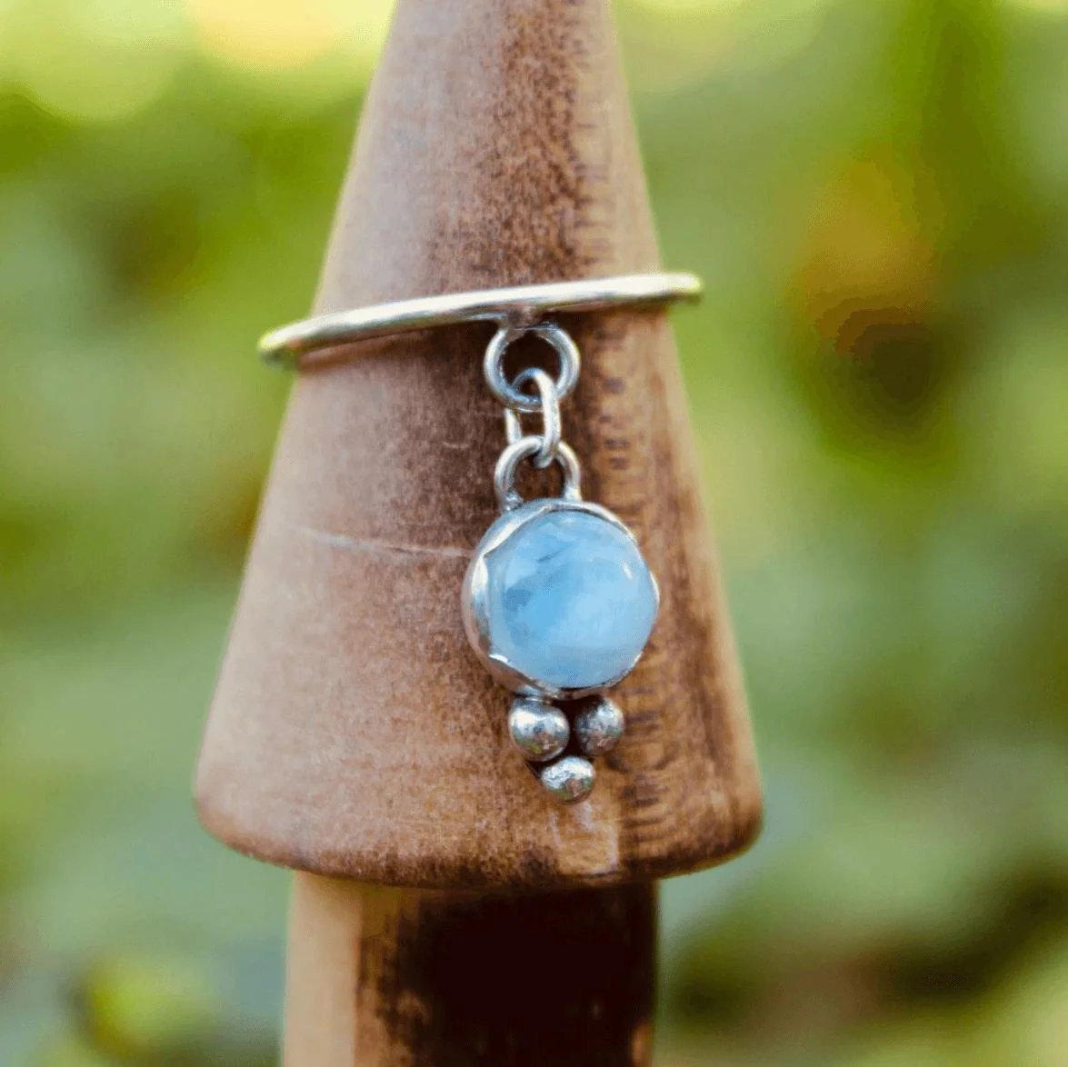 Close-up of a silver ring with a dangling blue gemstone and three silver beads, worn on a wooden cone-shaped object, with a blurred green and yellow background.
