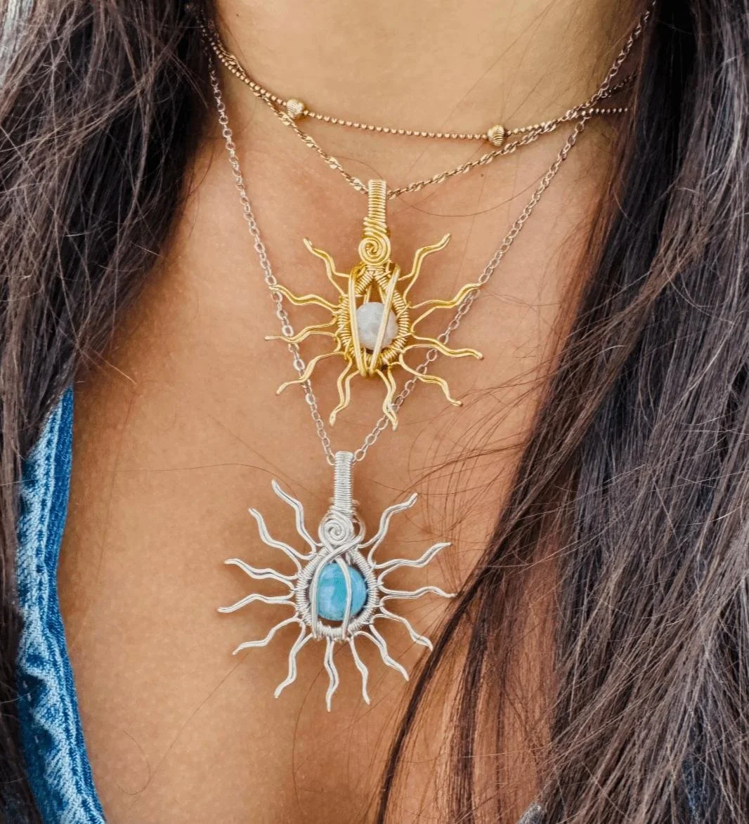 Three layered necklaces with sunburst designs; the top necklace is gold with a sun-like pendant with a white stone, the middle is gold with a sun-shaped pendant, and the bottom necklace is silver with a blue stone in a sunburst setting.