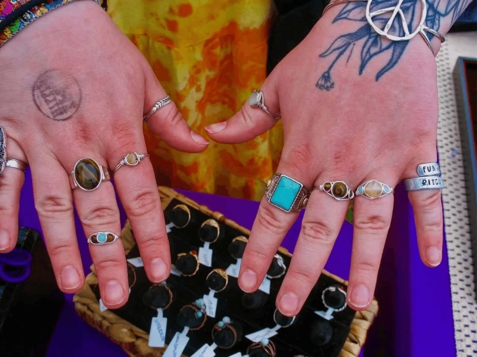 Person displaying multiple rings on their fingers, including a large turquoise ring, a silver ring with a blue stone, and a silver ring with the words 'FUN' and 'BITCH' inscribed. They have tattoos on their hands and are wearing a silver peace sign b