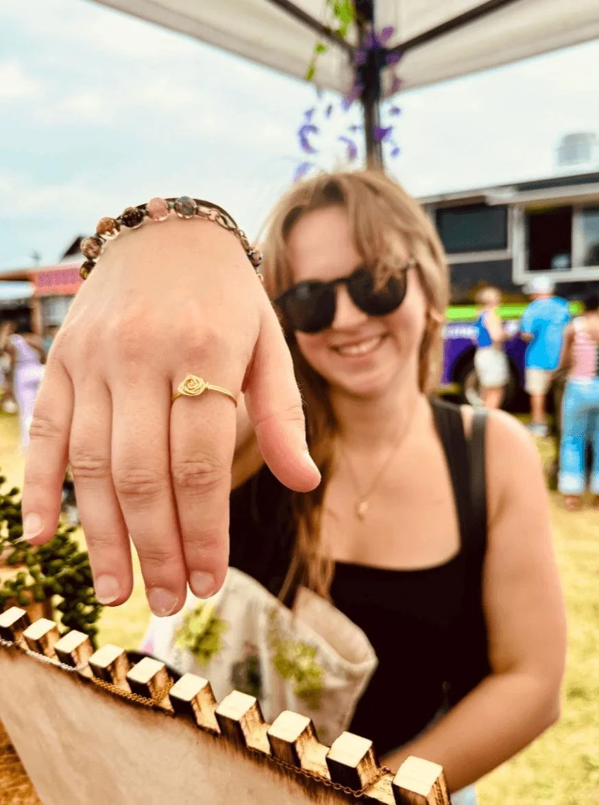 A smiling woman with sunglasses shows her hand with rings and jewelry while at an outdoor event, with a booth and other people in the background.