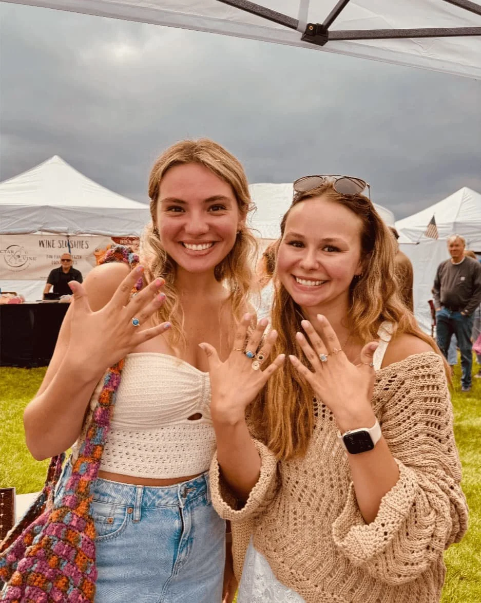 Two smiling young women showing off rings on their fingers at an outdoor event with white tents and gray sky in the background.