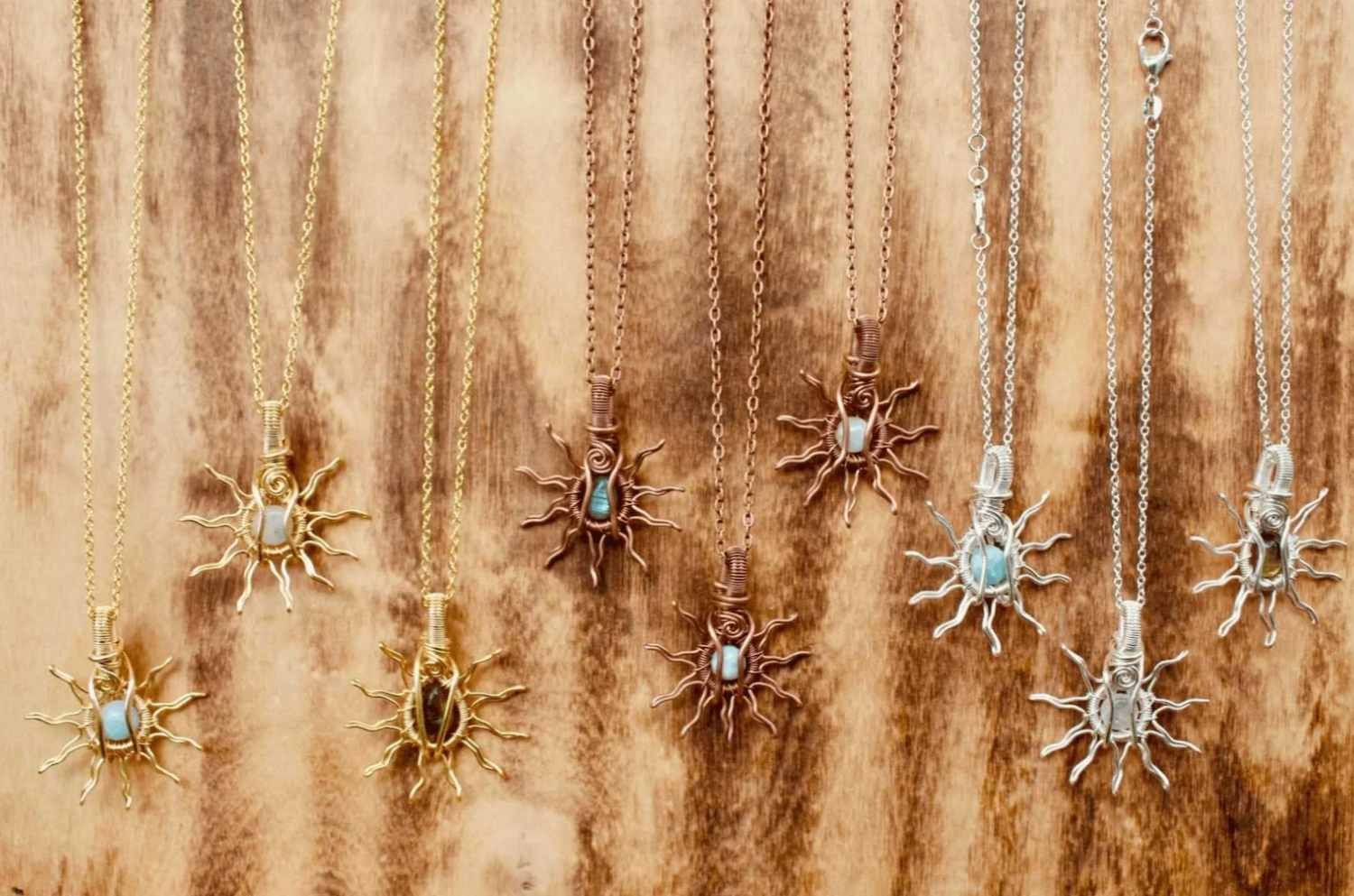 A display of ten necklaces hanging on a wooden wall, featuring leaf and organic motifs in gold, silver, and copper finishes, each with a central stone or bead.