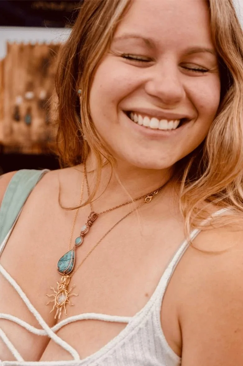 A smiling woman with reddish hair wearing layered necklaces, a cream-colored top with thin straps, and earrings.