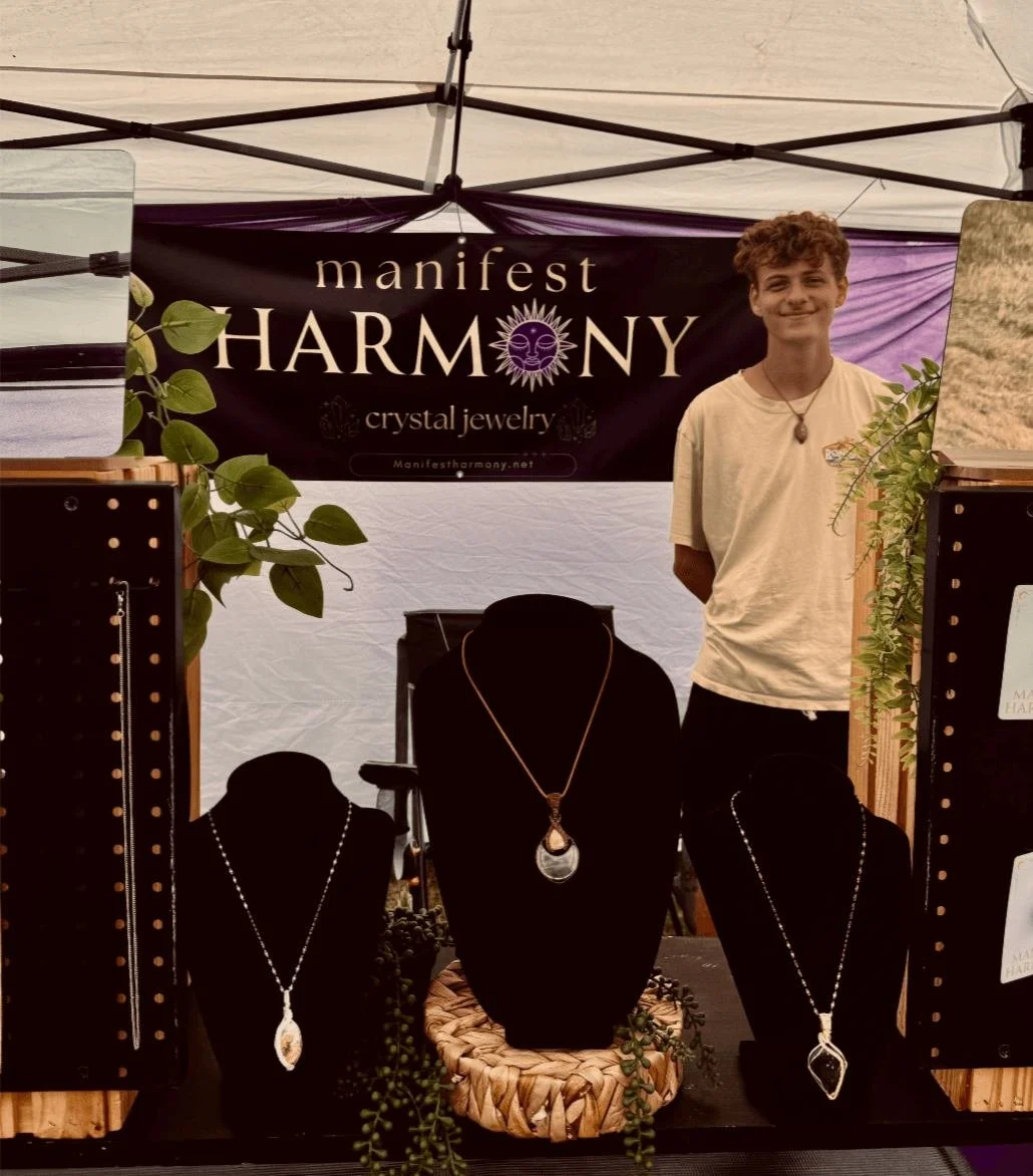 A young man standing behind a display of crystal jewelry at a market booth. The booth is decorated with green plants and has a banner reading "manifest HARMONY," with a website link. The jewelry includes necklaces with pendants arranged on black disp