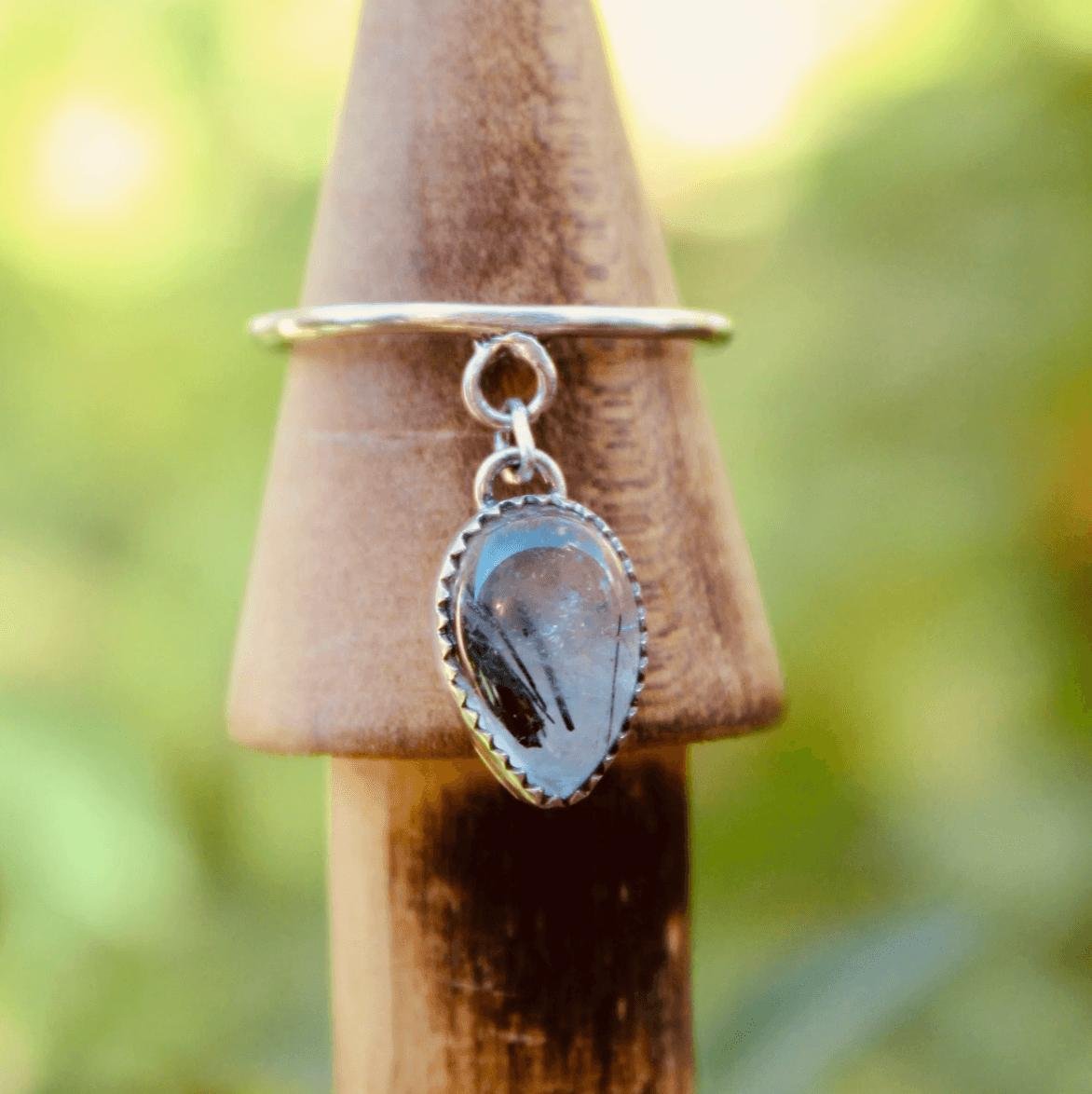 A silver ring with a dangling pendant featuring a polished black gemstone, hanging on a wooden cone called a ring holder, with a blurred green background.