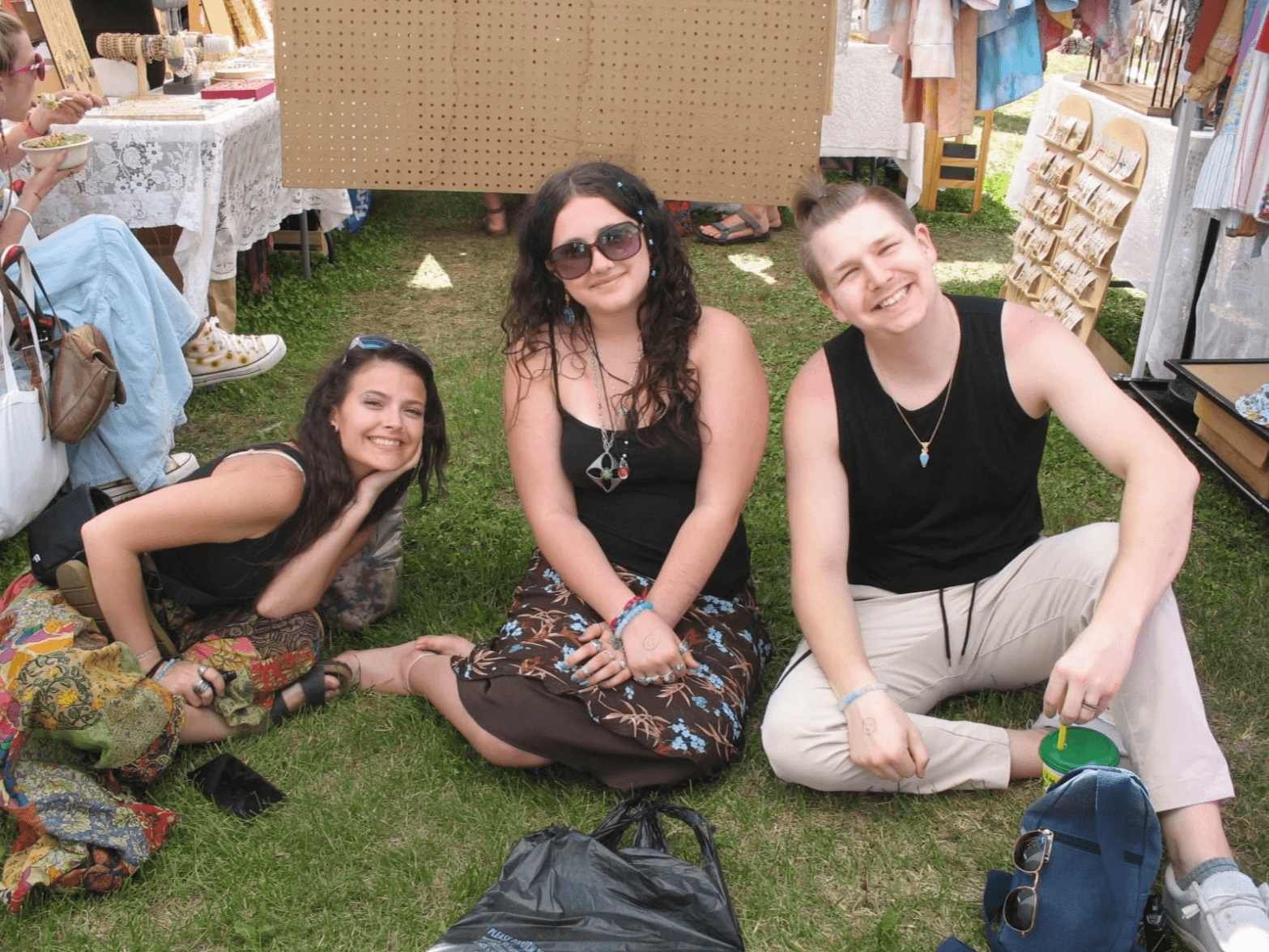 Three people sitting cross-legged on grass at an outdoor market or fair, smiling at the camera. The person on the left has long dark hair, wearing a black top and colorful patterned pants. The person in the middle has long curly hair, wearing sunglas