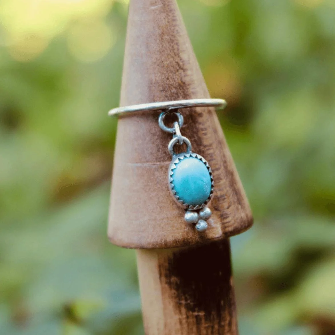 Close-up of a silver ring with a turquoise stone and dangling silver beads, mounted on a wooden cone-shaped stand with a blurred green background.
