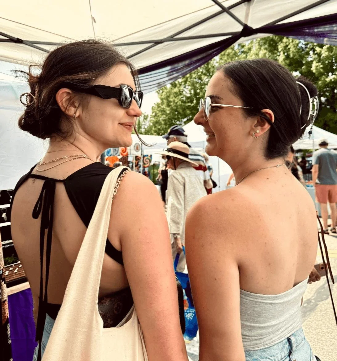 Two women with dark hair and sunglasses standing close together at an outdoor event, smiling at each other under a tent.