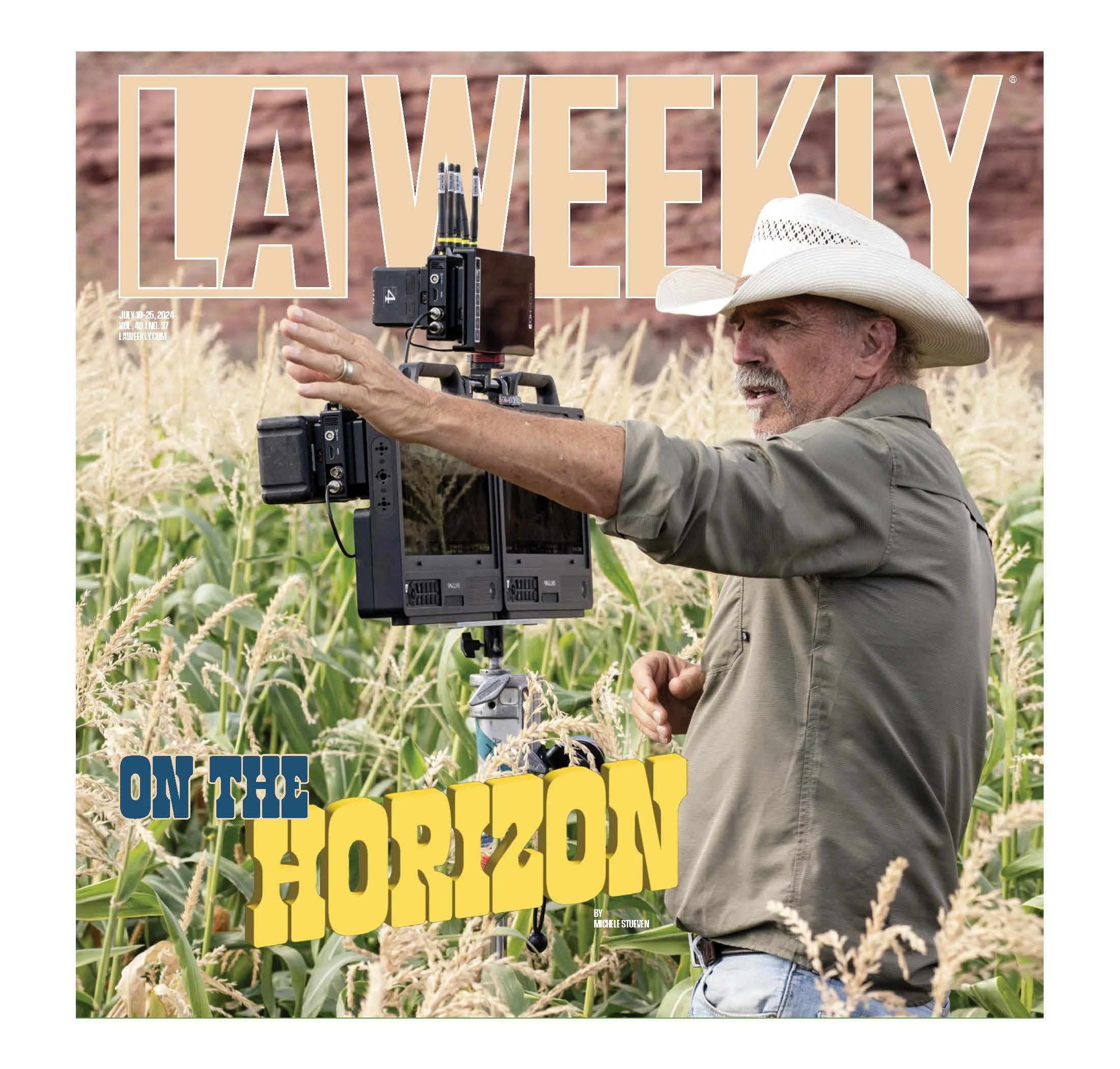 A man wearing a white cowboy hat and a khaki shirt operating a film camera on a tripod in a cornfield during daytime.
