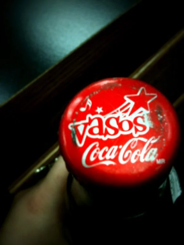 A Coca-Cola bottle with a red cap featuring the Vasos brand logo and festive star and musical note designs.