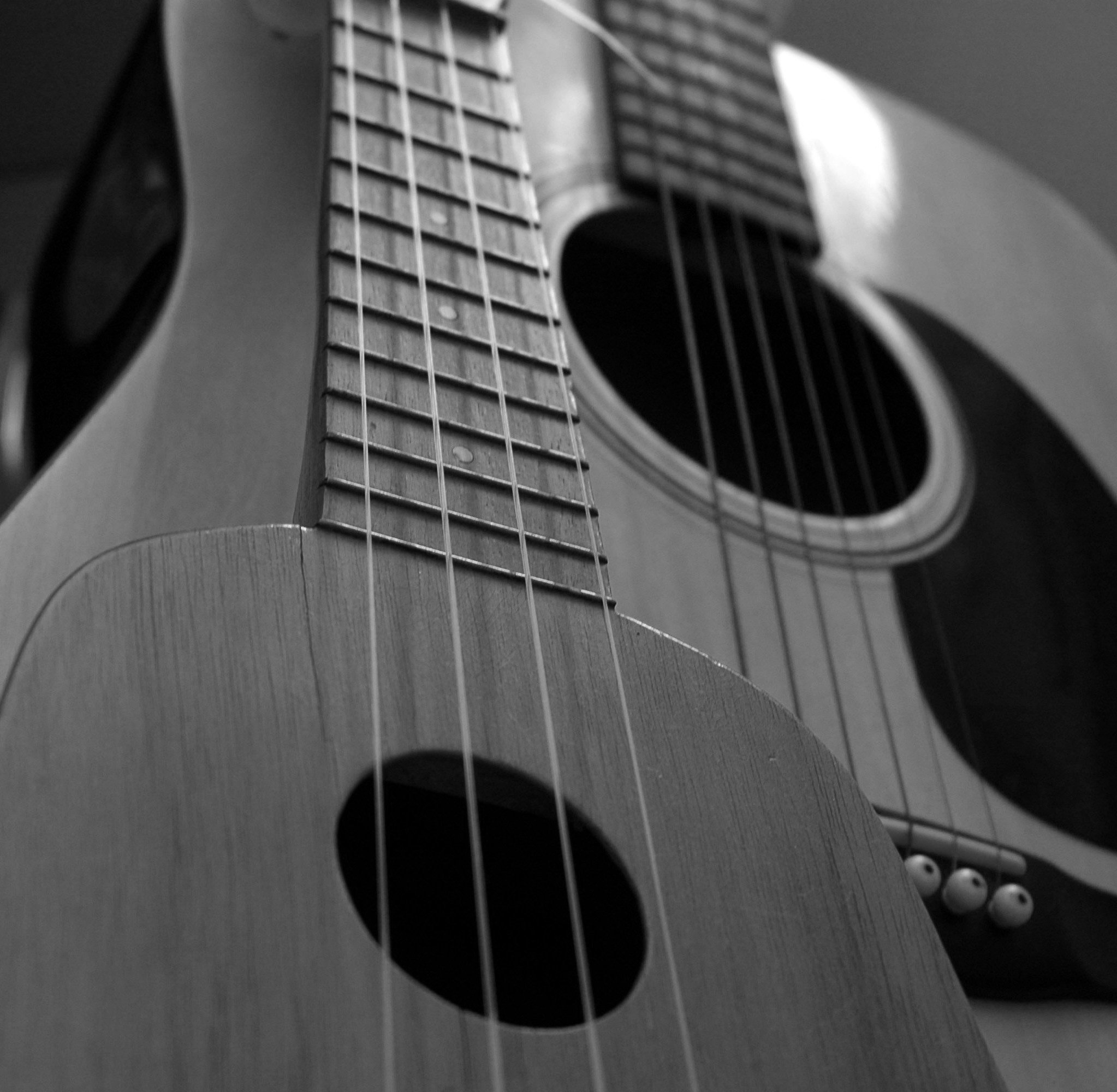 Close-up black and white image of an acoustic guitar's neck, strings, and sound hole, emphasizing the guitar's wood grain and string details.