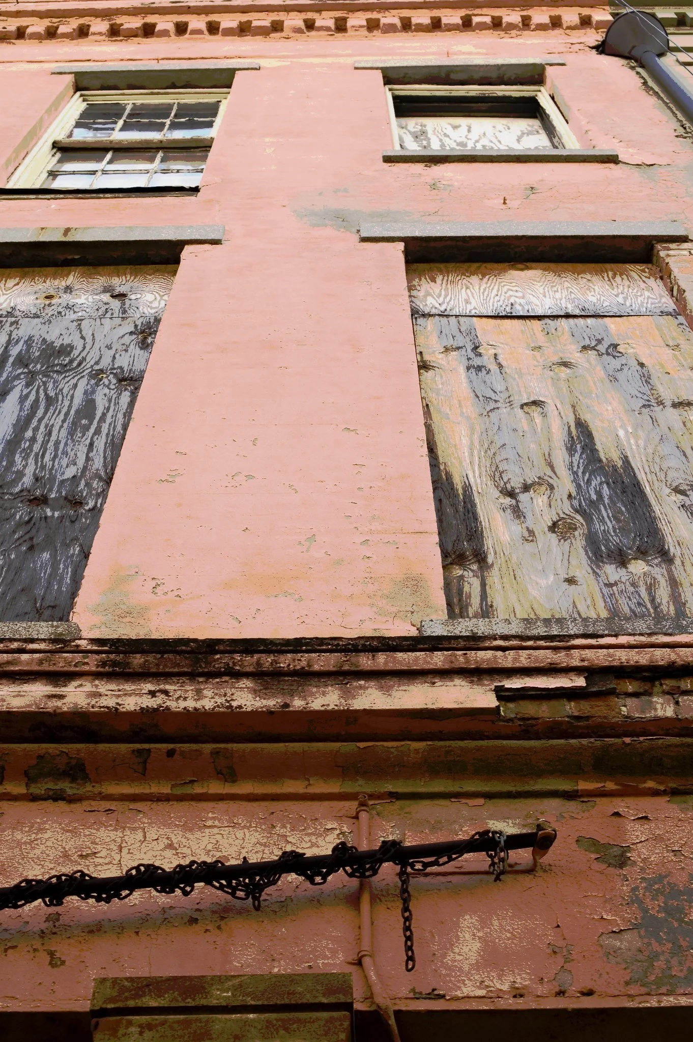 Close-up of an old, weathered brick building with peeling pink paint, boarded-up windows, and a chain-link fence.
