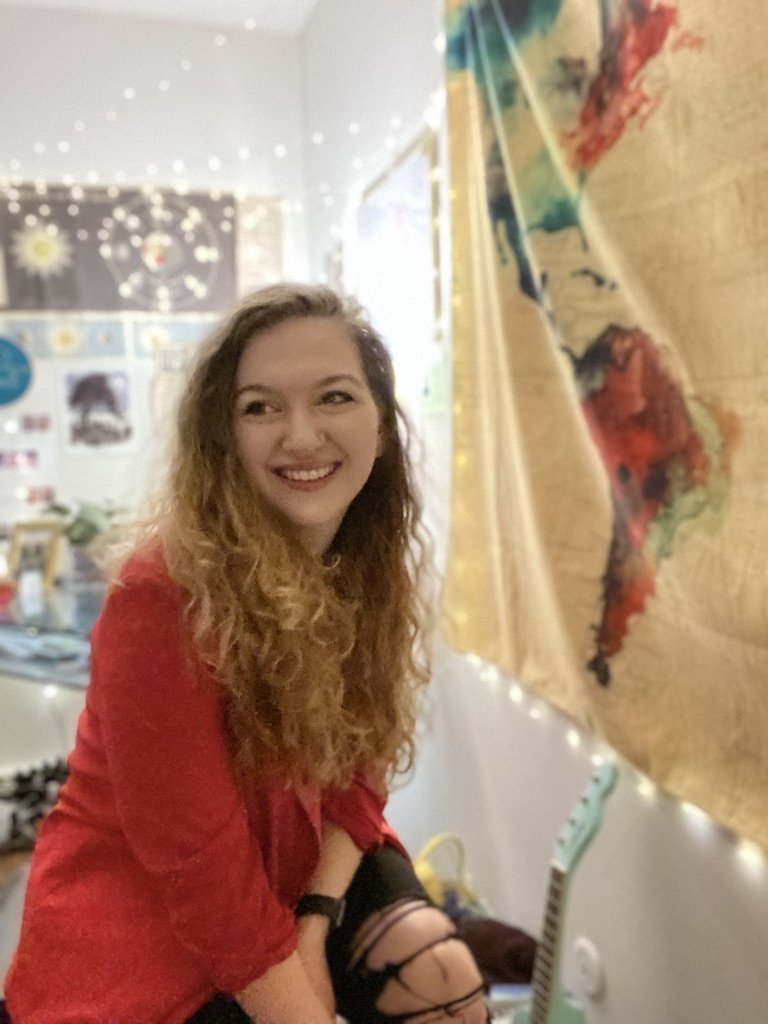 A young woman with curly hair smiling while sitting on a bed in a cozy, decorated room with string lights and a colorful map on the wall.