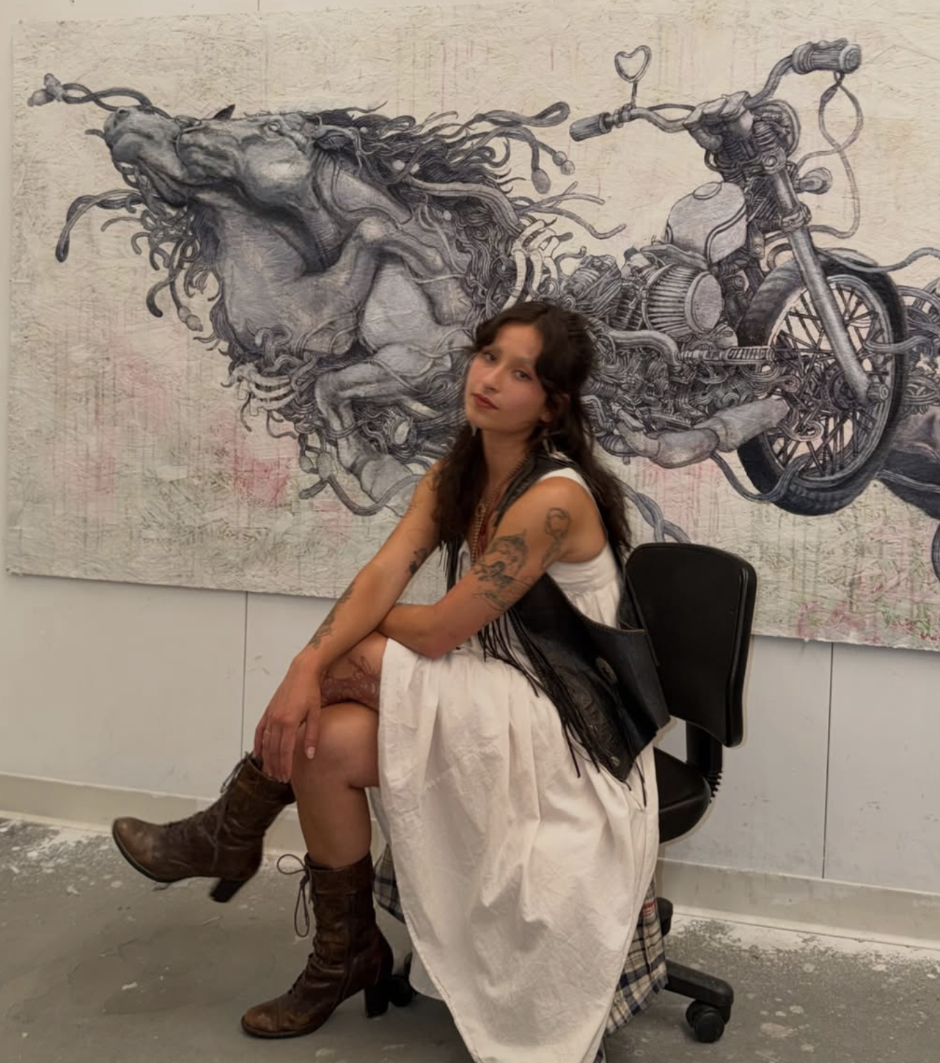 A woman sitting in a chair with her legs crossed, posing in front of a large, detailed mural of a horse and a motorcycle on the wall behind her.