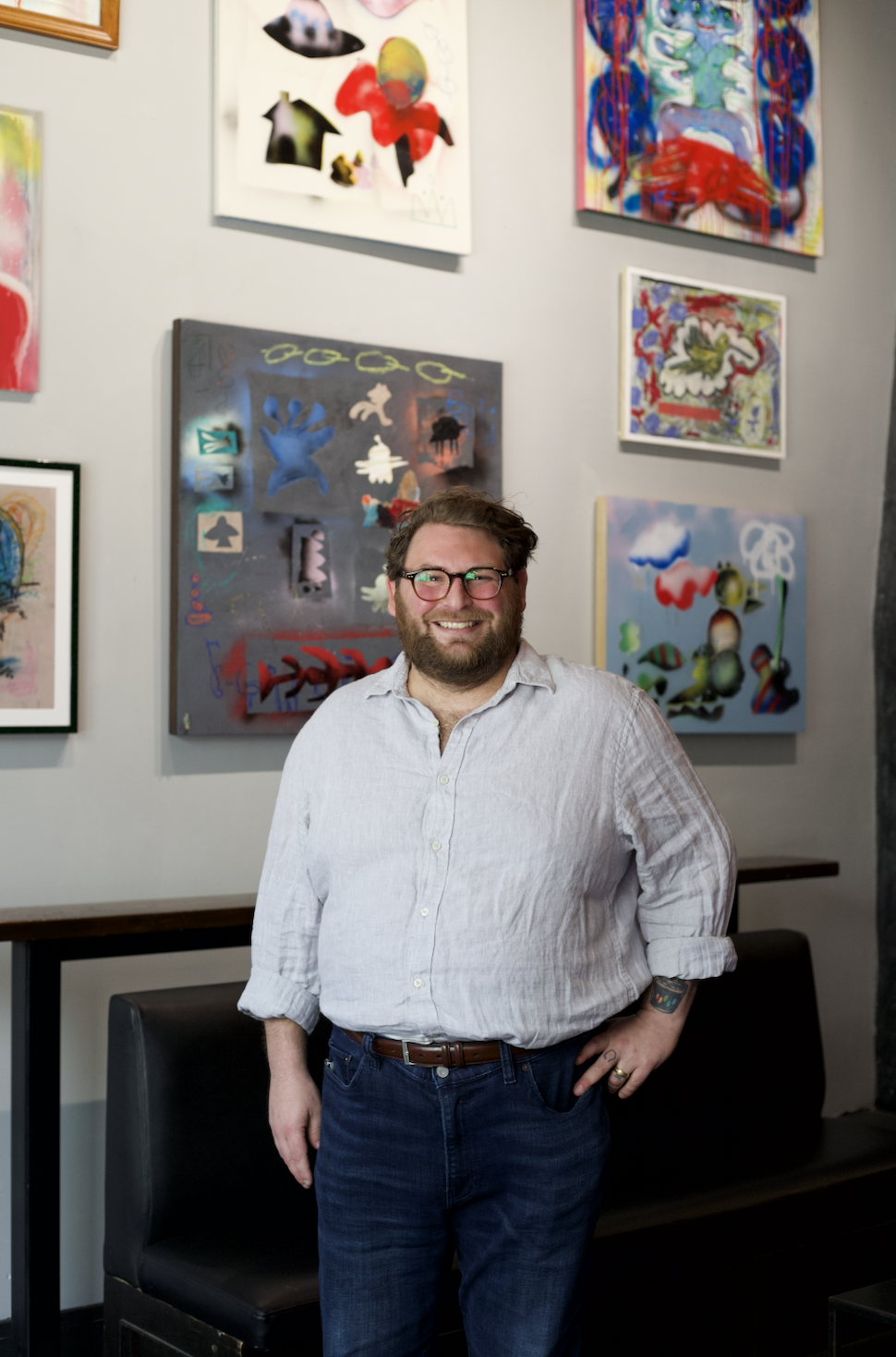 A man with glasses, a beard, and a white shirt smiling while standing in front of a wall decorated with colorful abstract paintings at an art gallery or exhibition.