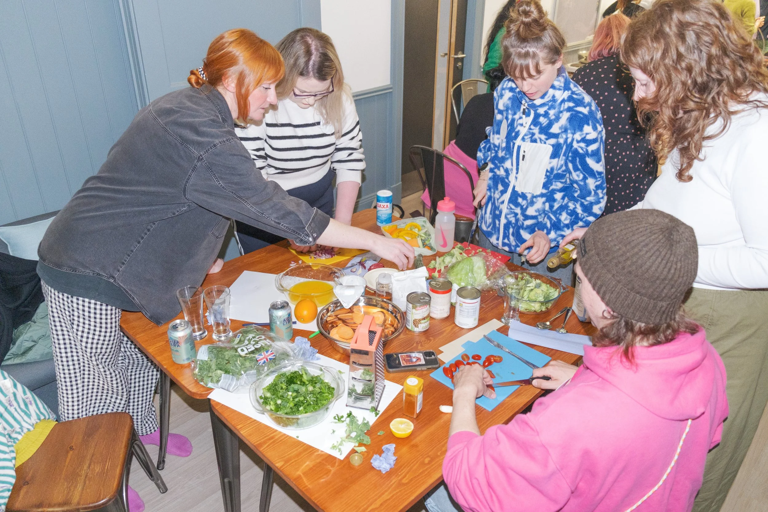 Group of people cooking and preparing food on a table with various ingredients like vegetables and canned goods.