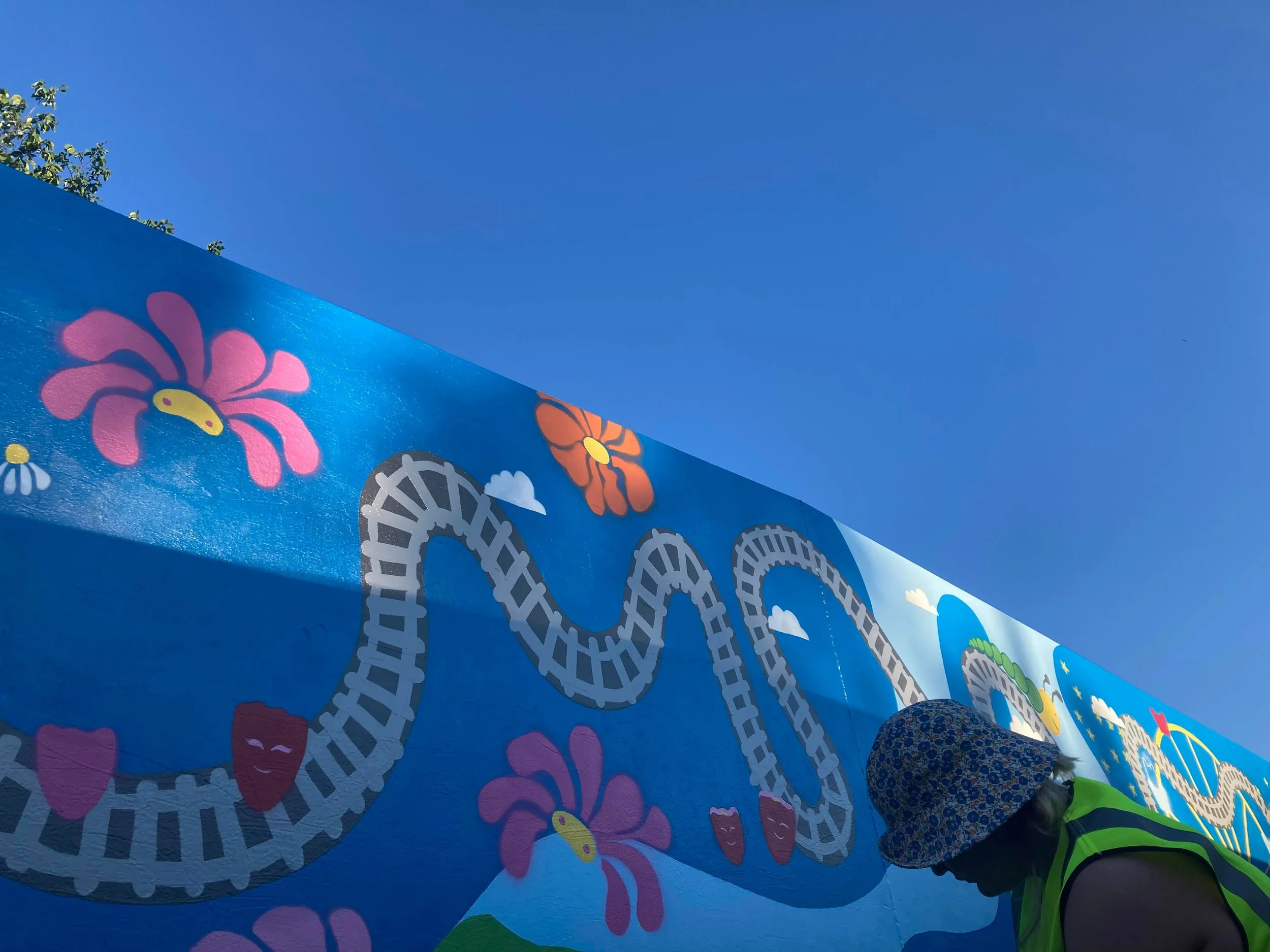 Mural with vibrant colors, featuring a winding train track, large flowers, and clouds against a bright blue sky. A person in a floral hat and green vest is visible in the foreground.