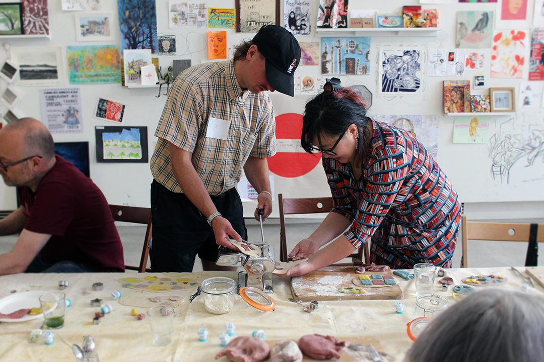 Two people working together at a crafting table, using a pasta machine. The room has artwork and sketches on the walls. A table in front is cluttered with crafting materials, jars, and utensils.