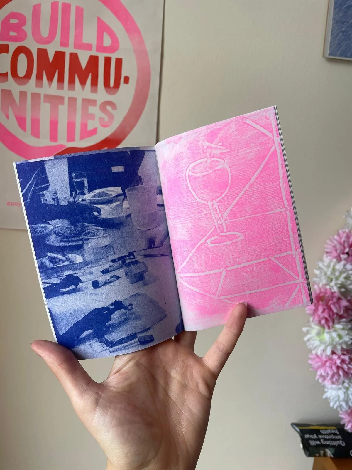 Person holding a booklet with colorful pages; left page in blue showing an abstract scene, right page in pink with a line drawing. In the background, a poster partially showing the words 'Build Communities.' Floral decoration on the right.