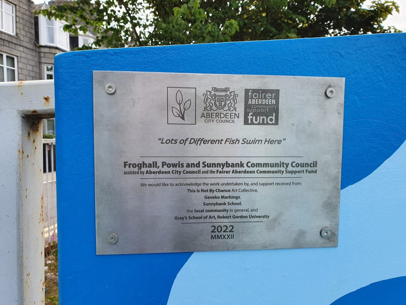 Plaque on a blue panel with text acknowledging a community project supported by Aberdeen City Council and Fairer Aberdeen Fund, involving various contributors, dated 2022.