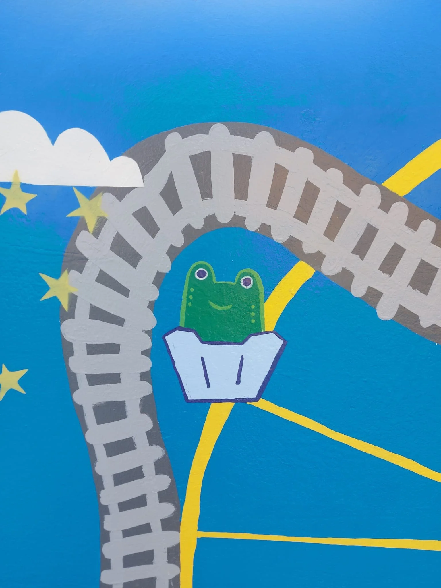 Colorful mural featuring a cartoon frog on a roller coaster, with stars and clouds in the background. The track is grey with a yellow path, set against a bright blue sky.