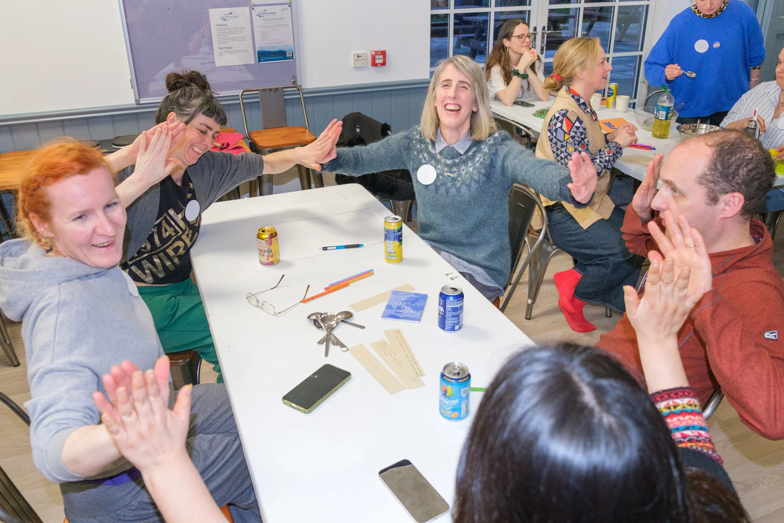 Group of people sitting around tables in a classroom setting, engaged in a lively activity, with drinks, papers, and pens on the table.