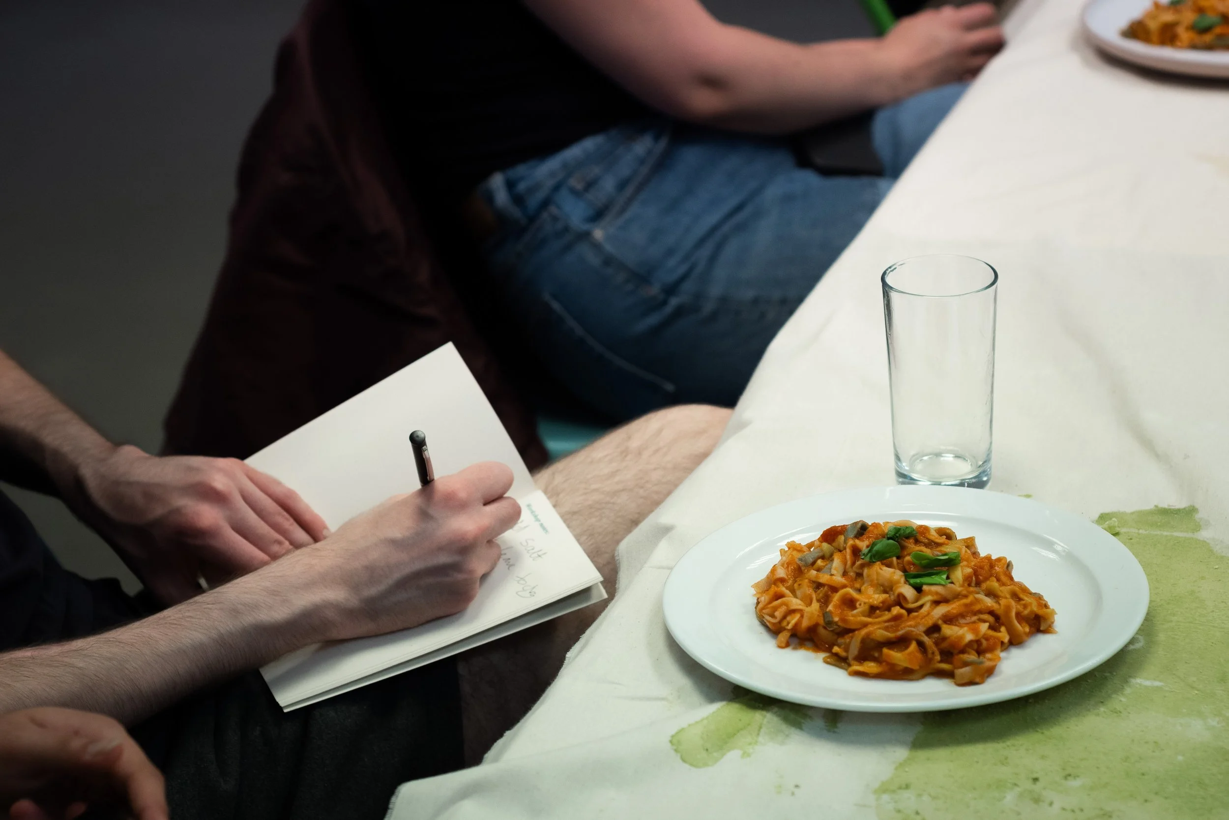 A person writing in a notebook with a pen while sitting at a table with a plate of pasta and an empty glass. The tablecloth is stained green.