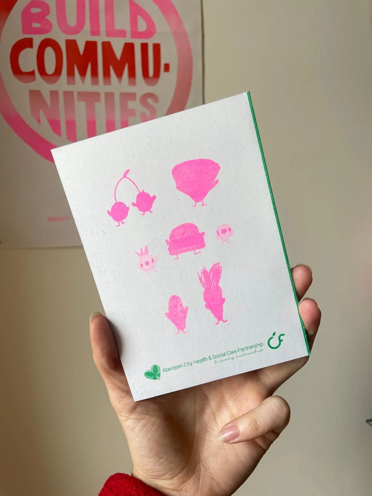 A hand holding a card with pink cartoon animal illustrations. The card has text at the bottom reading 'Aberdeen City Health & Social Care Partnership.' In the background, red text on a poster reads 'Build Communities.'