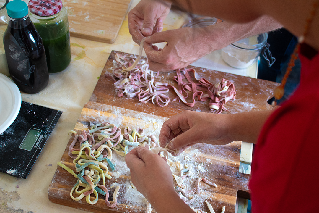 People making homemade colorful pasta on wooden cutting boards, surrounded by flour, bottles, and kitchen tools.