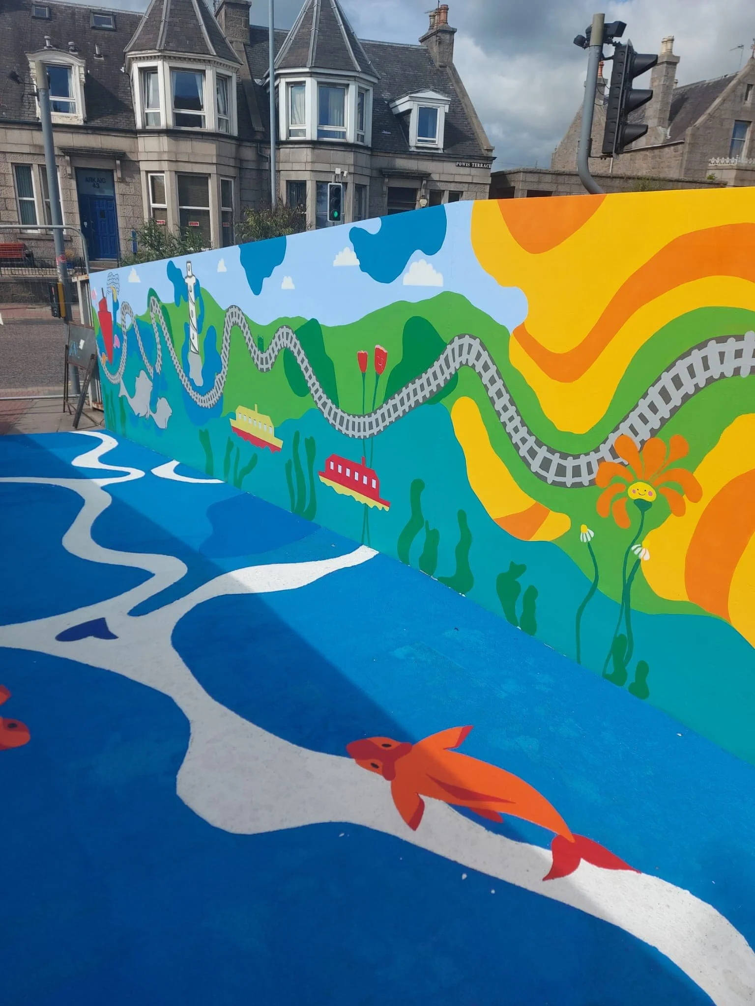 Colorful mural featuring train tracks, landscapes, and bright elements with a blue ground depicting a river and cartoon-like fish. Urban background with stone buildings and traffic lights.