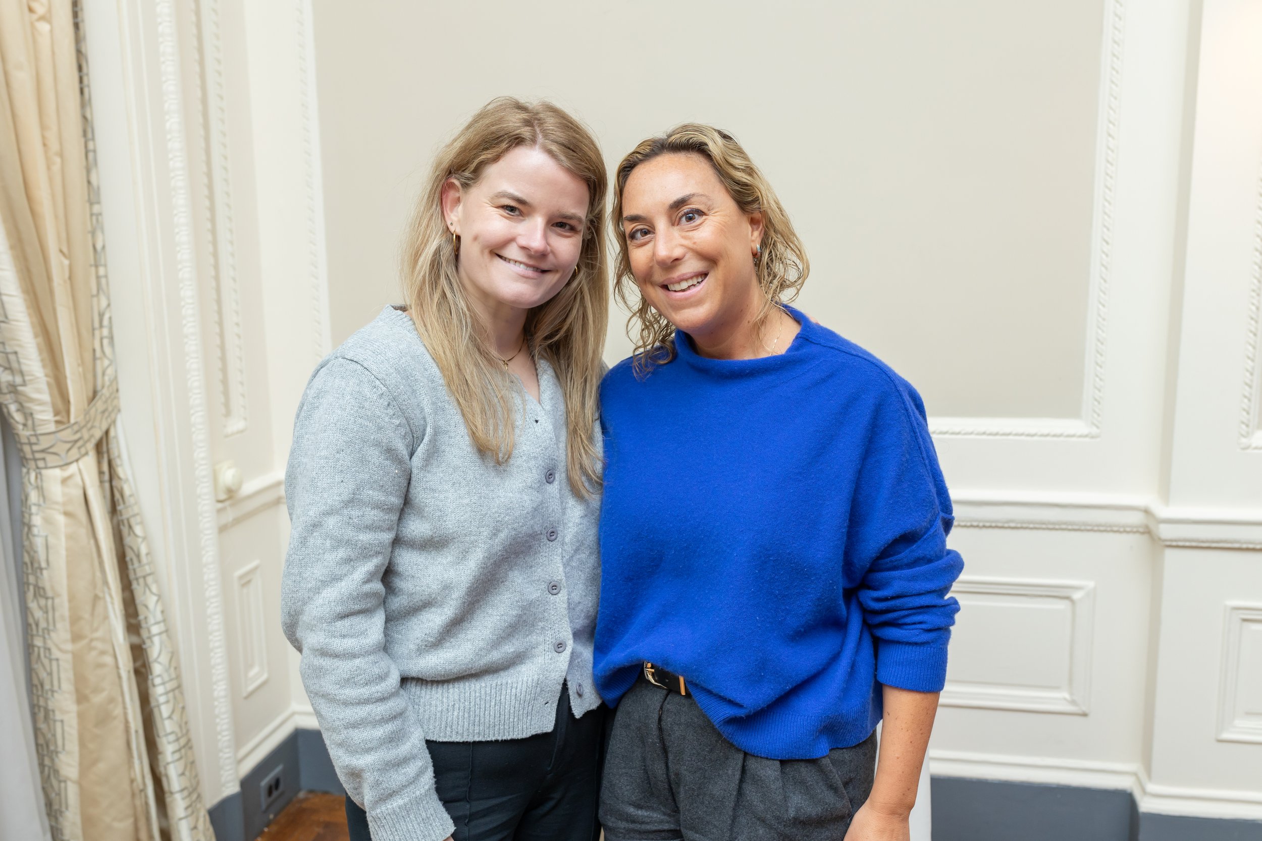 Two women smiling and posing in a room with white paneled walls and beige curtains, one wearing a gray sweater and the other a blue sweater.