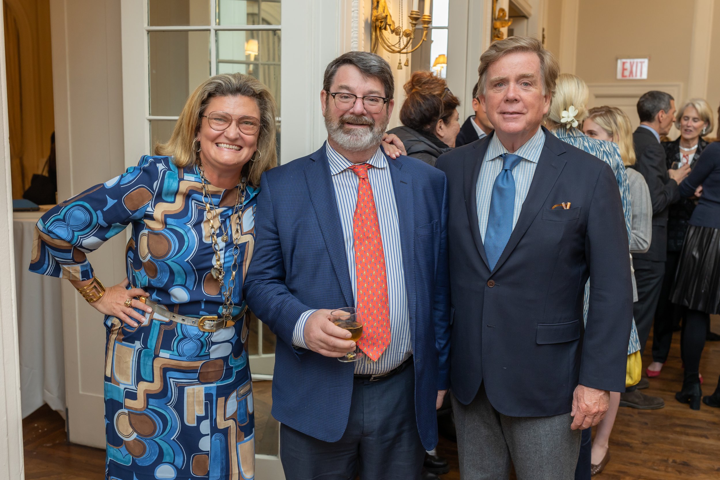 Three people posing together at a social event, one woman in a patterned dress, and two men in suits, one holding a drink. In the background, more people are socializing.