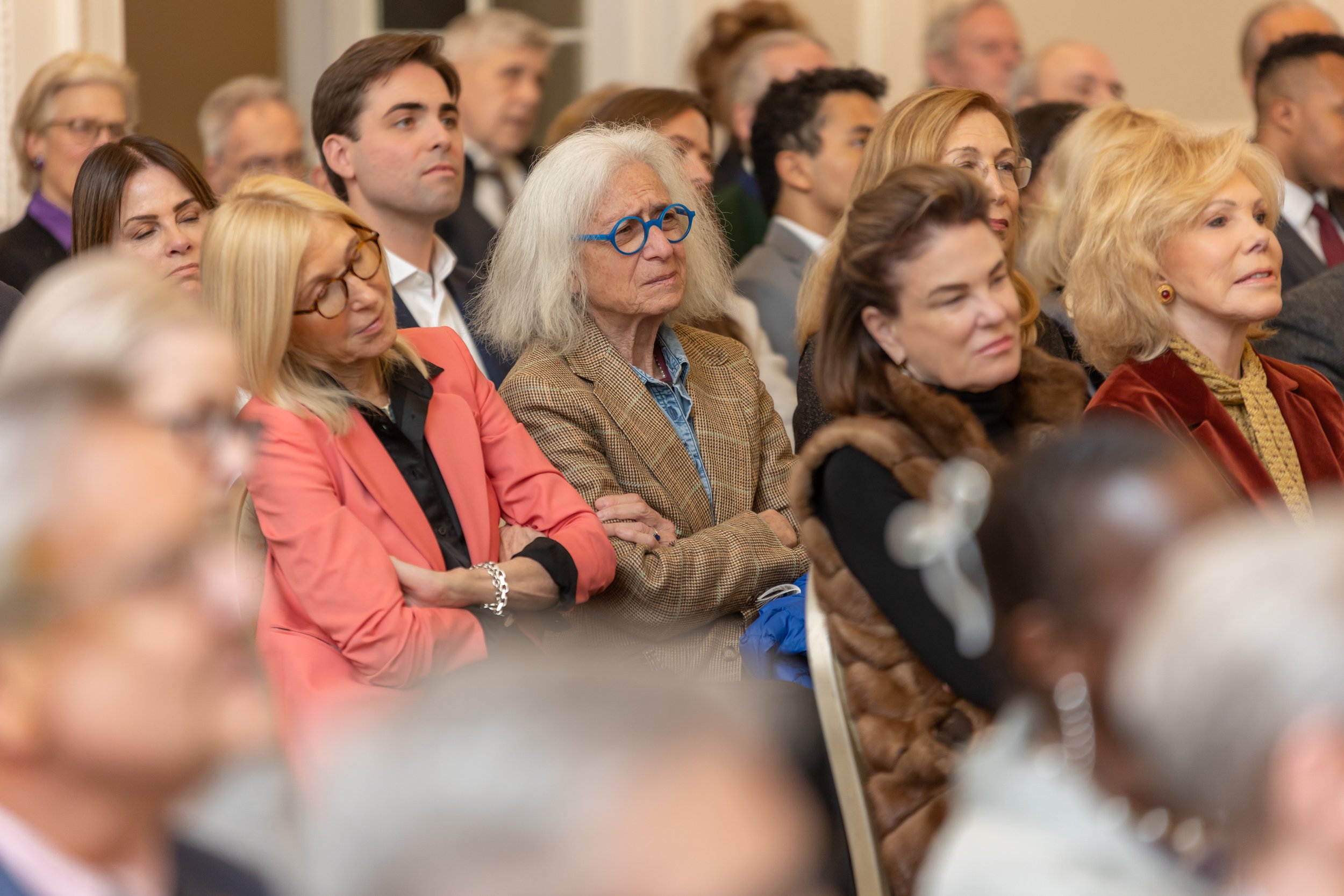 A group of diverse people seated, attentively listening during an indoor event. Various ages and styles are represented, including a woman with white hair and blue glasses wearing a checkered blazer.