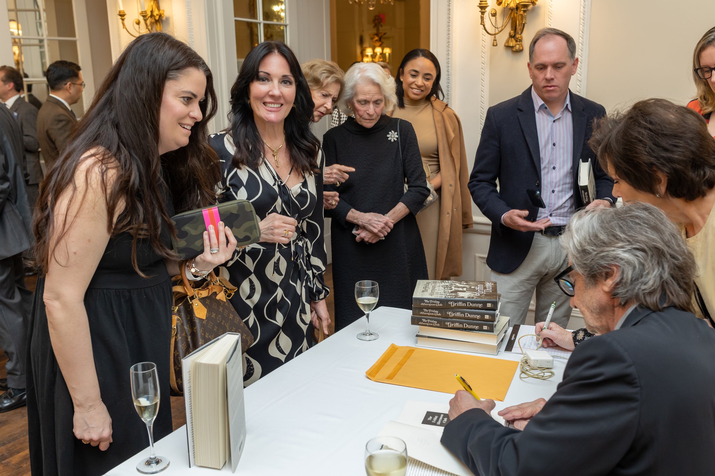 People at a book signing event with an author signing books, surrounded by attendees holding books and talking.