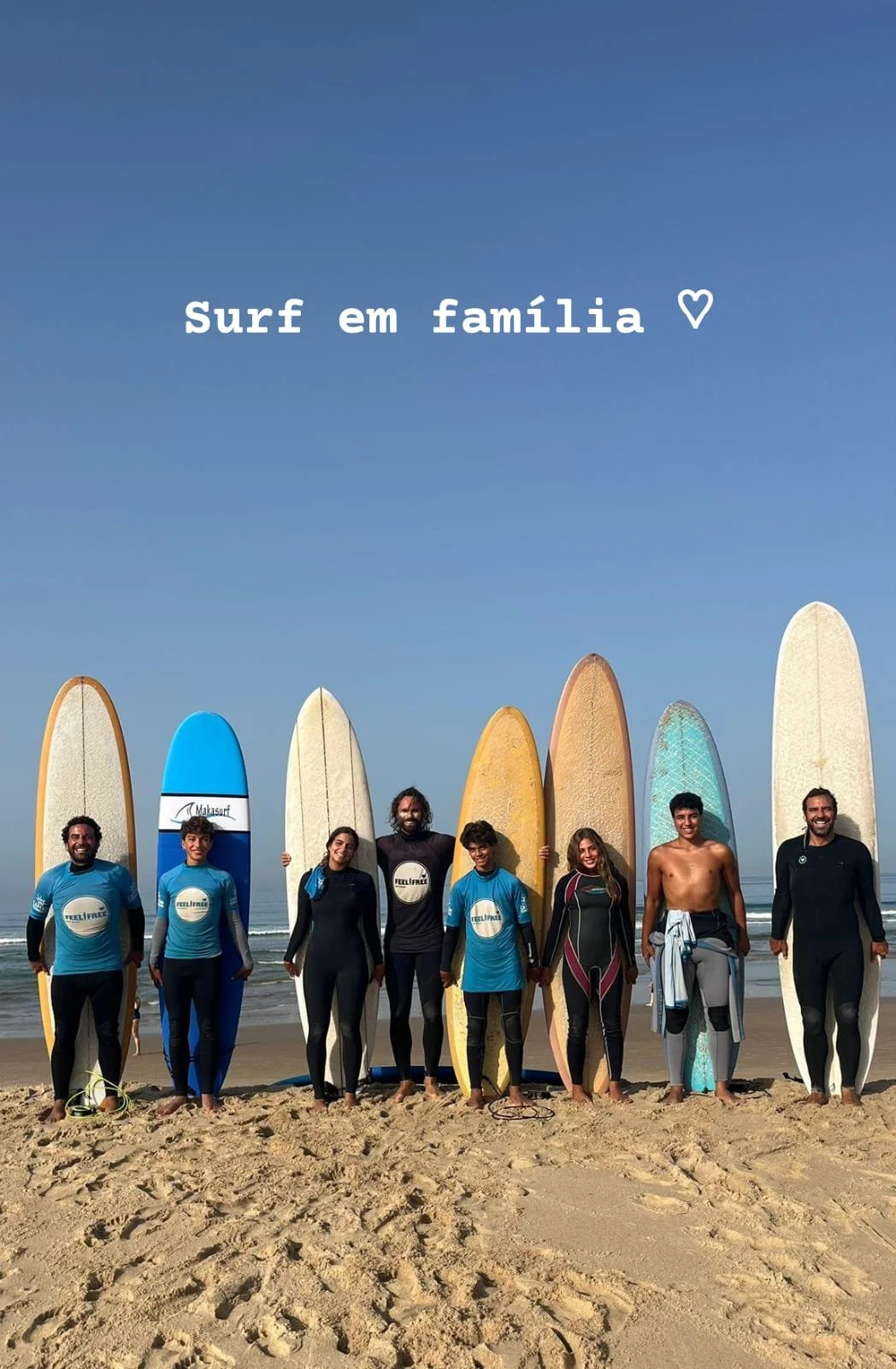 Family of eight people having a surf class together on a beach holding surfboards, smiling, with text 'Surf em família' and a heart symbol at the top.