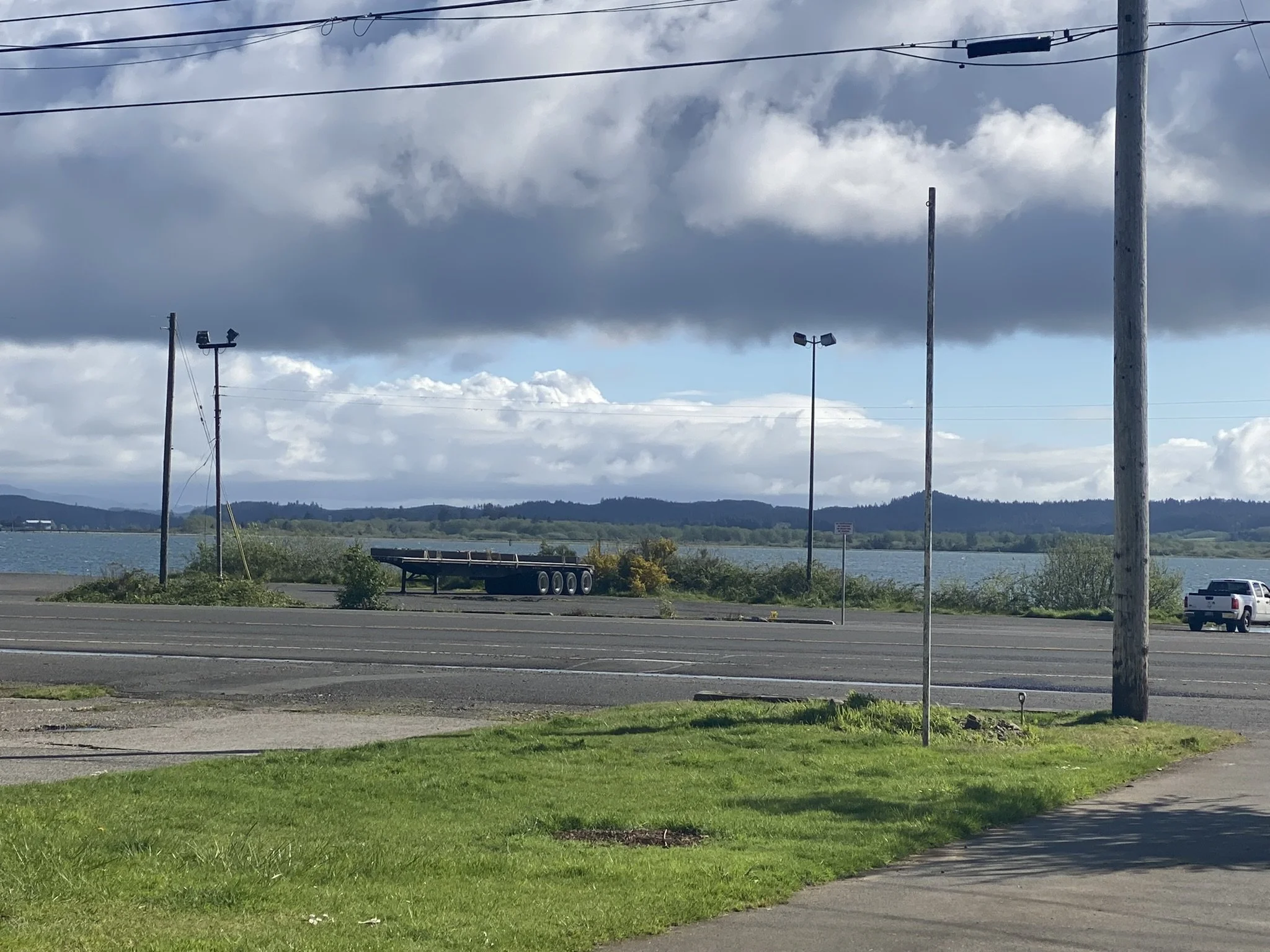 A parking lot next to a body of water with distant hills and cloudy sky. Utility poles and wires are overhead, and a few vehicles are parked or driving.