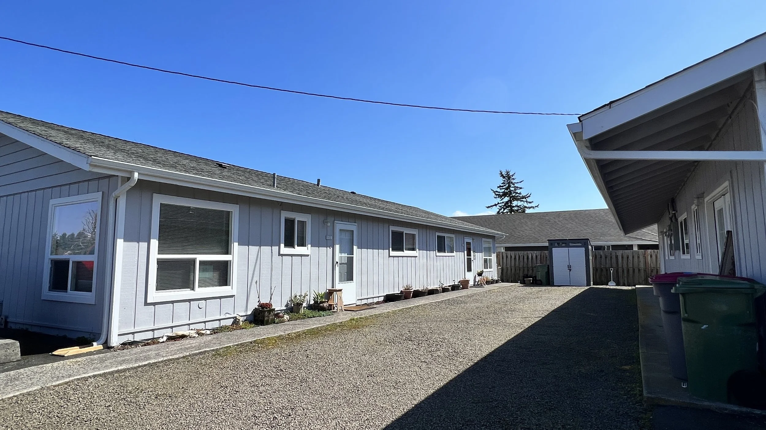 Backyard with light gray house, gravel driveway, potted plants, shed, and trash bins, under a bright blue sky.