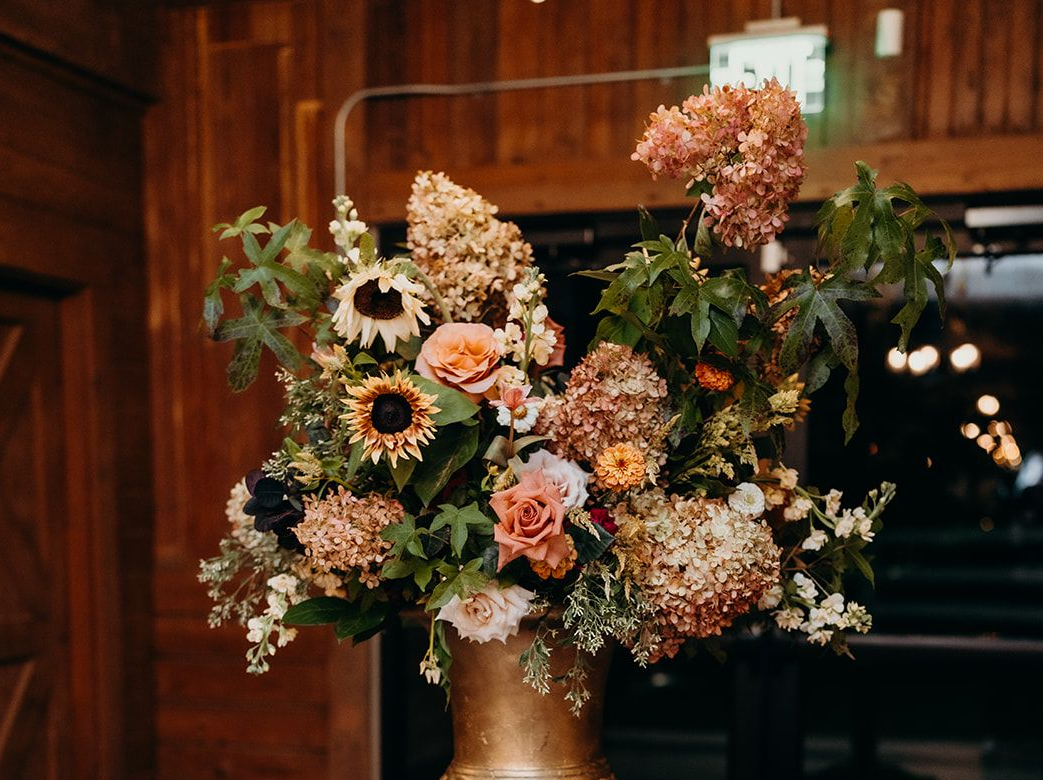 Lush floral arrangement with autumn blooms of sunflowers, hydrangea, and roses in a gold large vase for an entrance statement