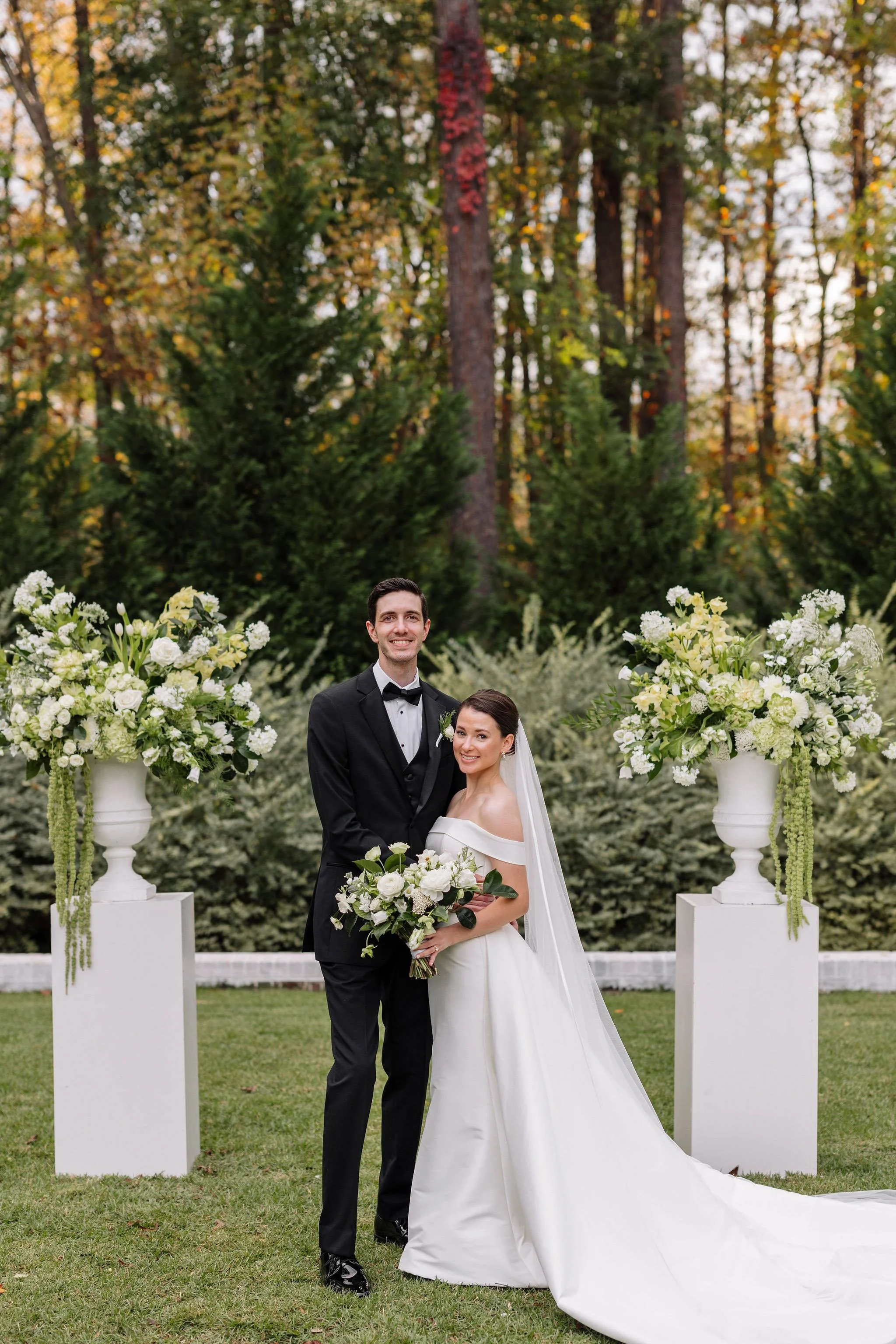 Bride and groom stand in front of two pedestal flower arrangements in white and green with hanging amaranthus