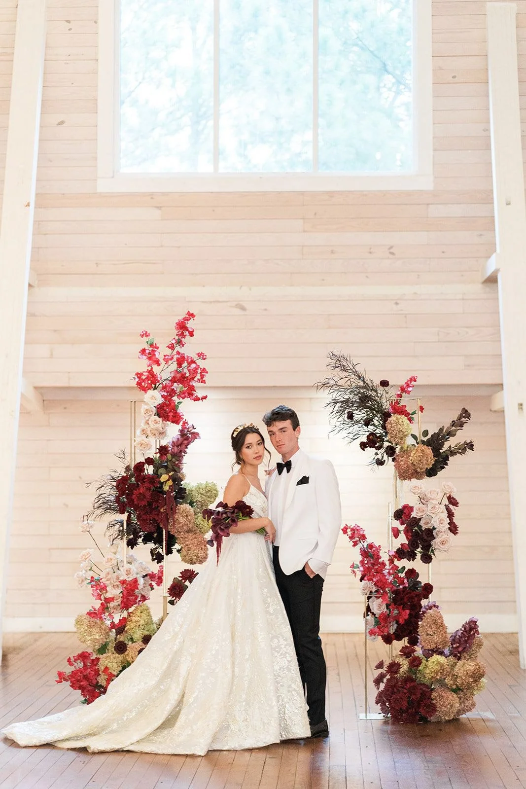 Bride and groom standing between dramatic floral pillars designed with deep red, blush, and neutral blooms at Historic Wakefield Barn, showcasing statement wedding florals that frame the ceremony space.